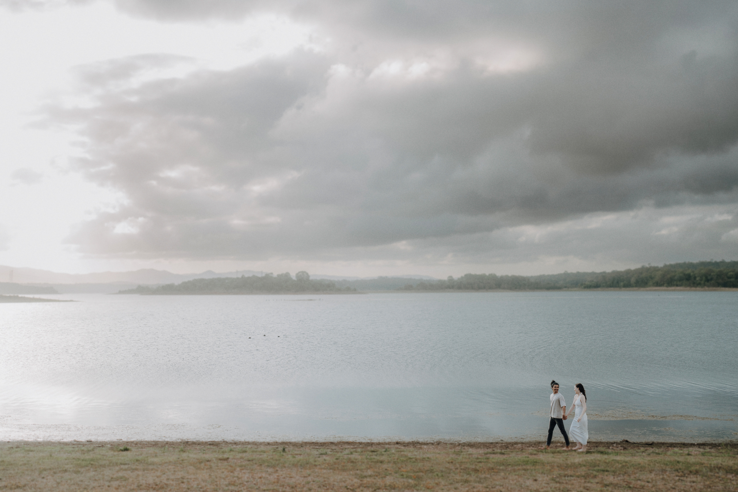 A couple walks along the grassy shore of a large lake under a cloudy sky, with distant hills and calm water in the background.