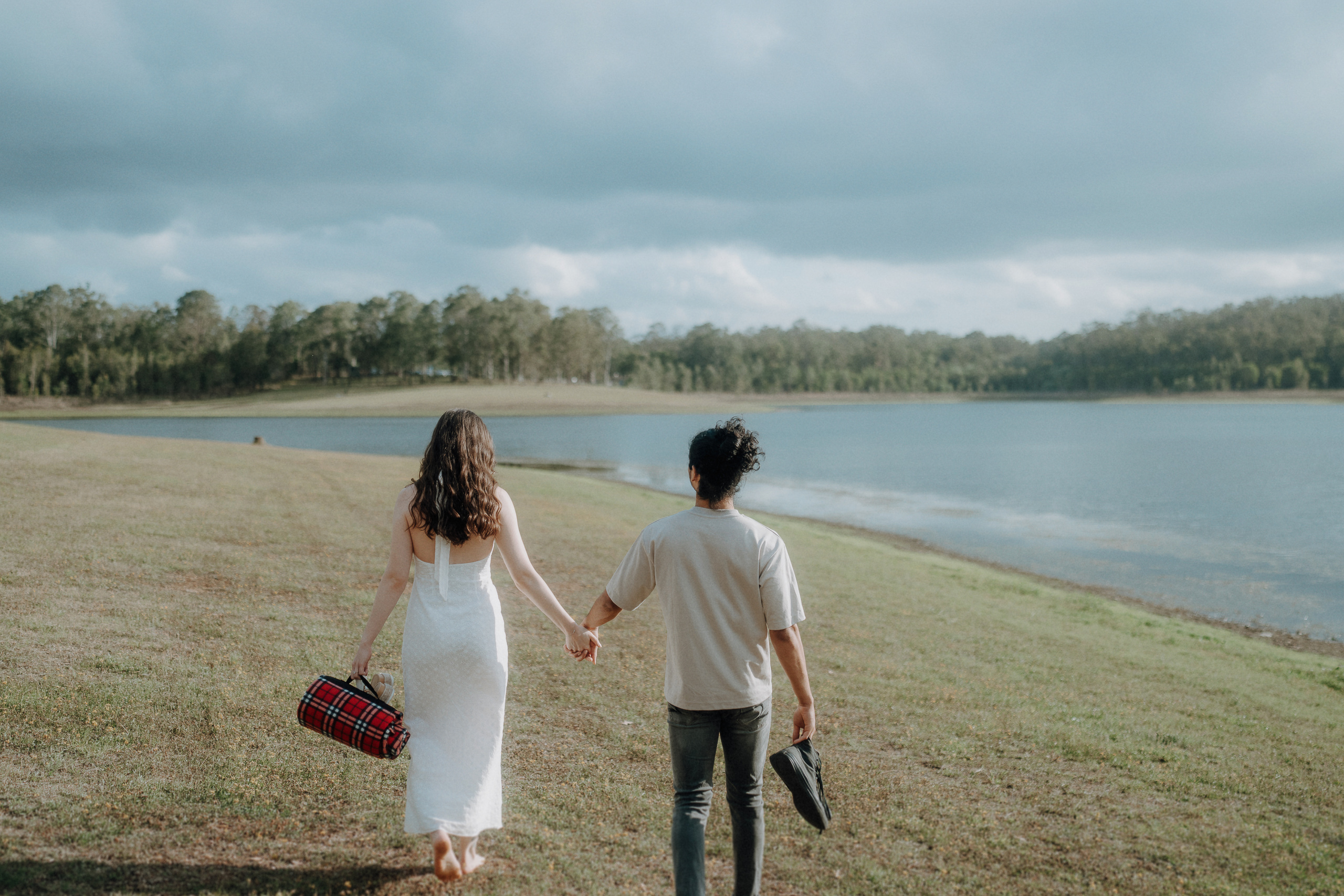 Two people walk hand in hand along a grassy lakeshore. One carries a plaid picnic basket, and the other holds shoes. Trees and water are visible in the background under a cloudy sky.
