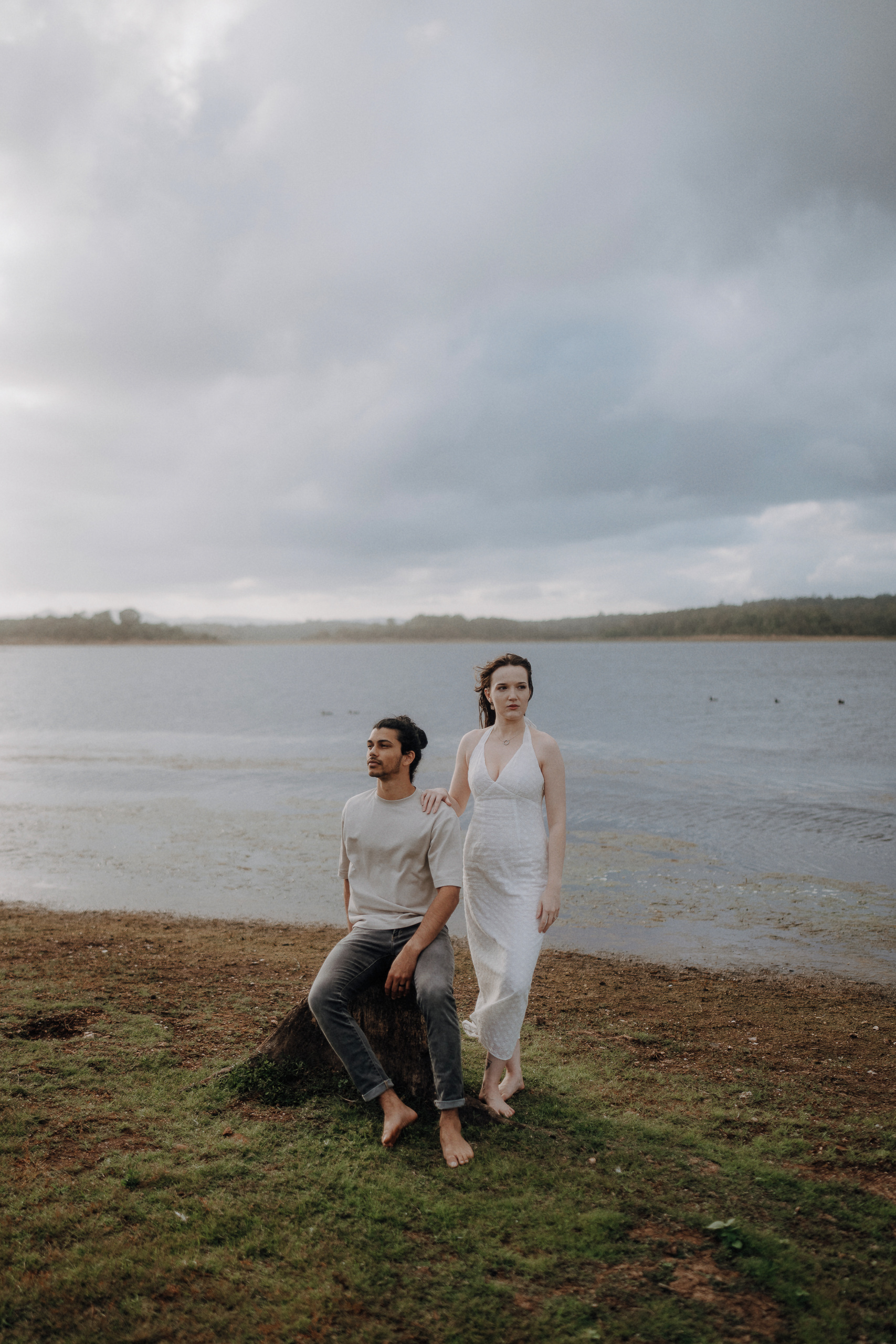 A man sits on a rock by a lakeshore while a woman in a white dress stands beside him, both barefoot, under a cloudy sky.