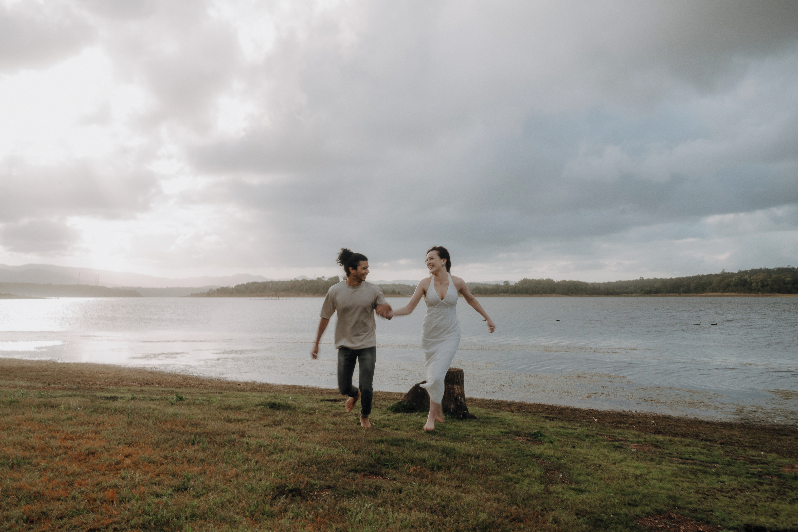 Two people hold hands and walk barefoot on grass along the edge of a lake under a cloudy sky.