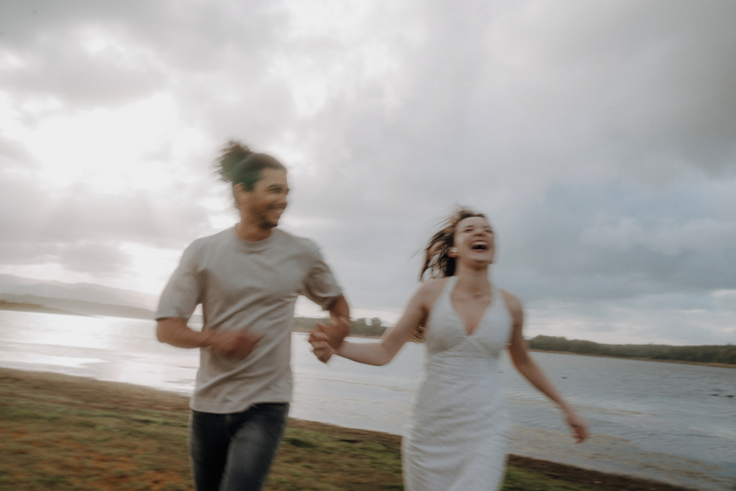 Two people holding hands run joyfully along a lakeshore, with water and cloudy sky in the background.