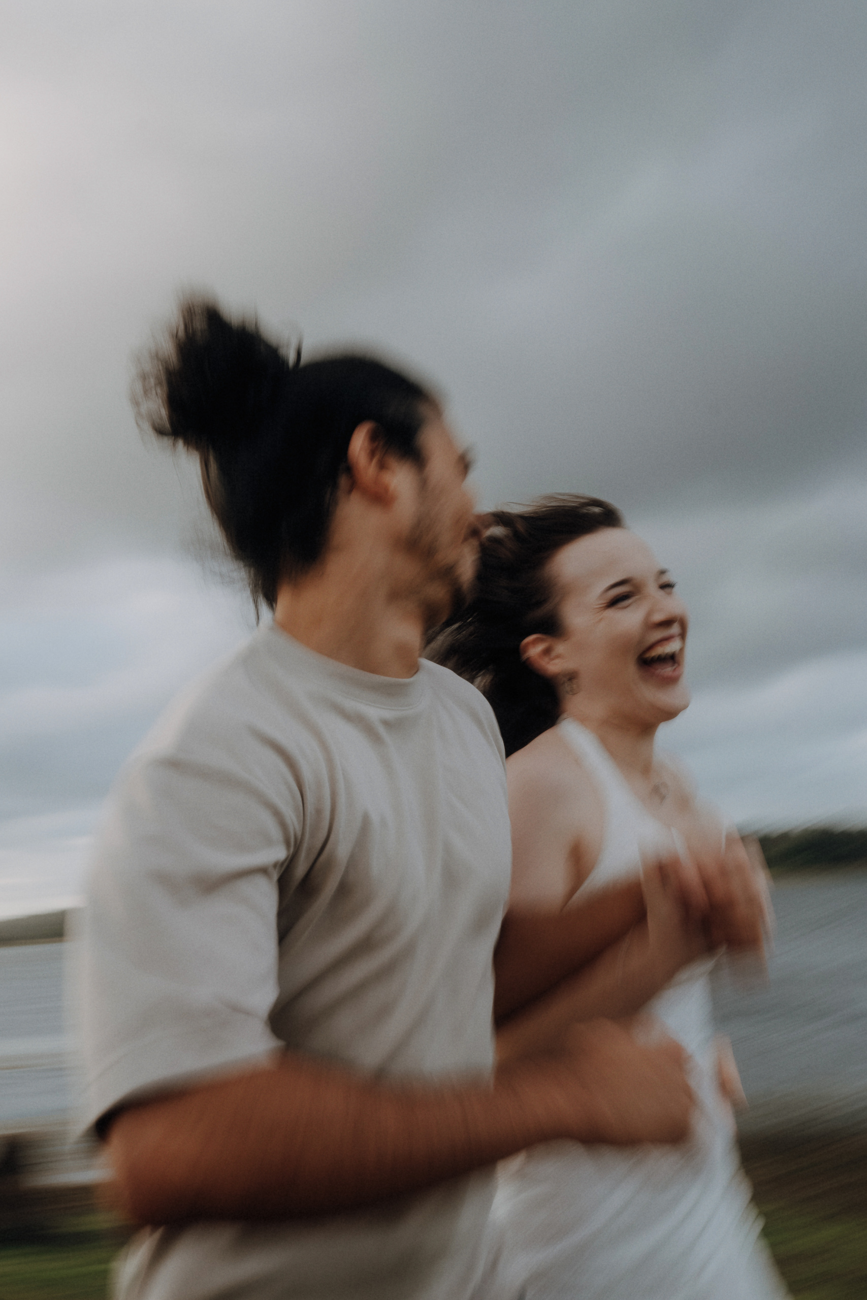 Two people holding hands and running outdoors near water, both smiling and captured in motion with a blurred background.