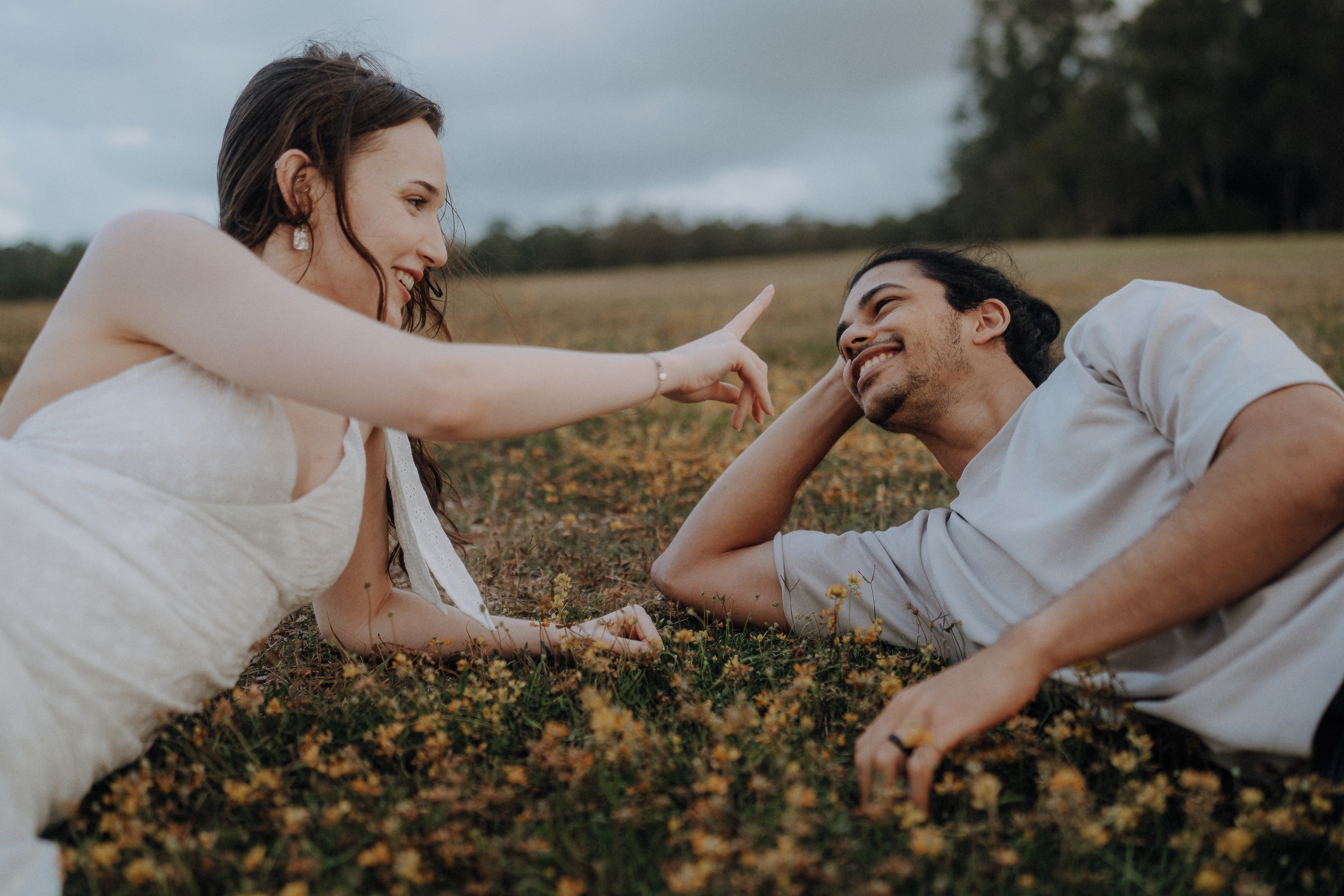 A woman in a white dress and a man in a light shirt lie on grass facing each other, smiling, as the woman playfully points at the man’s face.