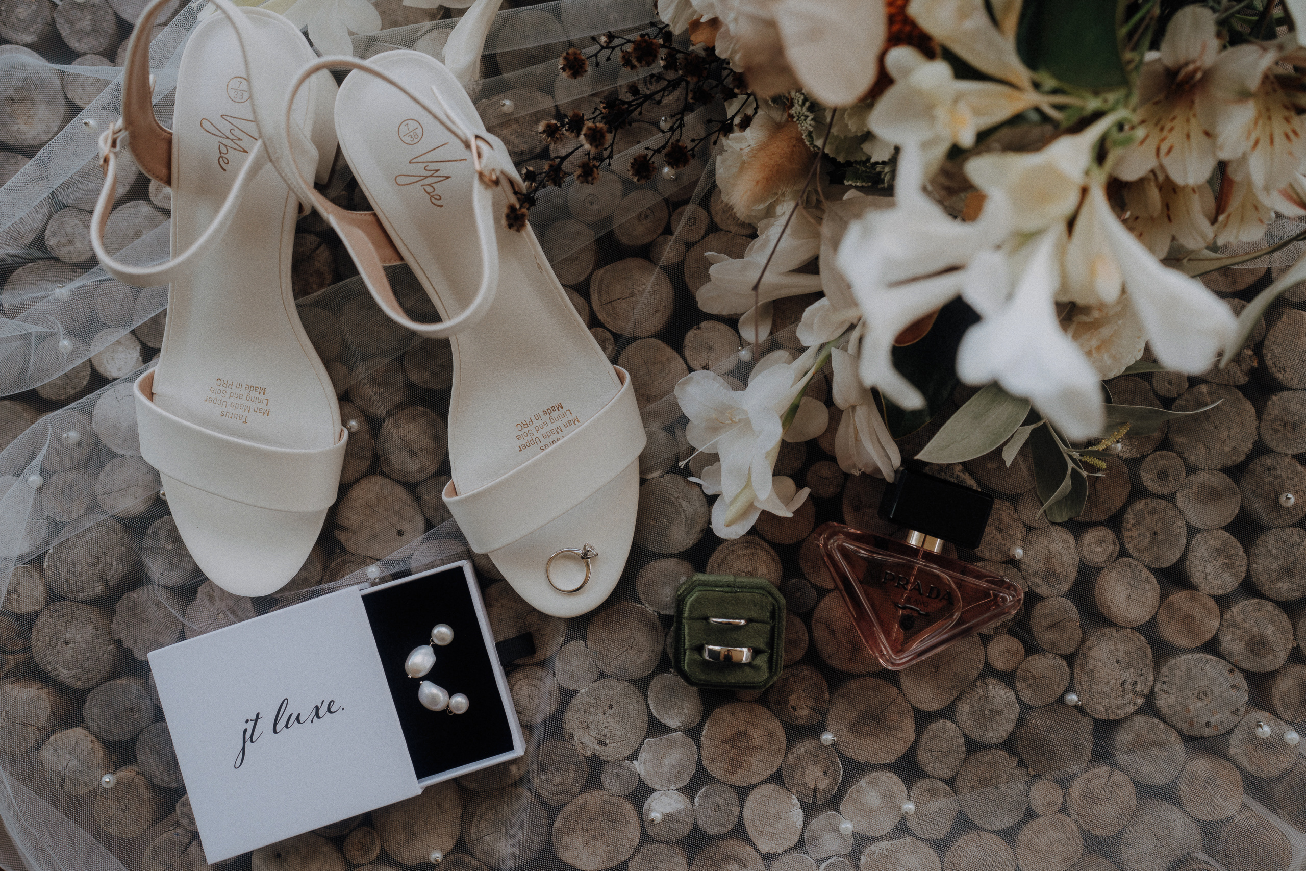 A pair of white heeled sandals, a jewelry box with pearl earrings, a gold ring, a green velvet ring box, a perfume bottle, and a bouquet of white flowers on a wooden surface.