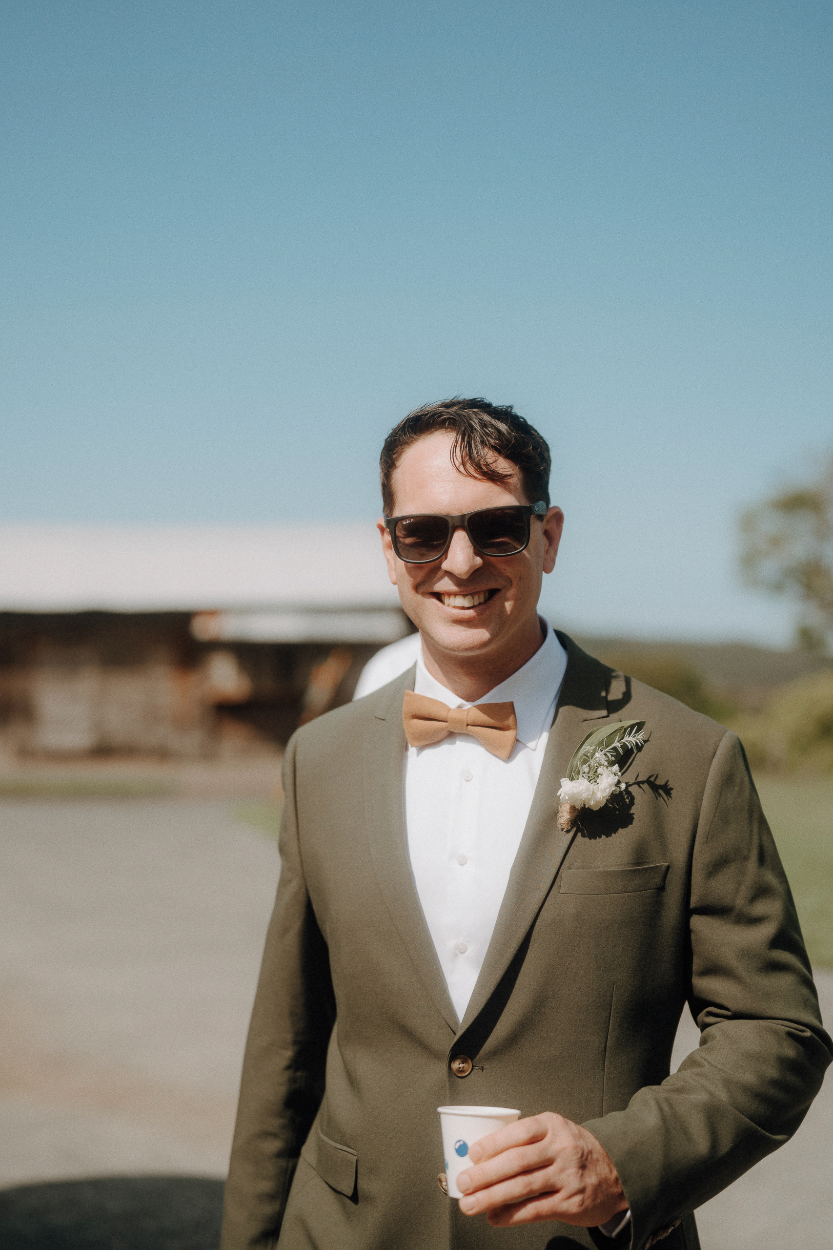 A man in a green suit with a bow tie and boutonniere stands outdoors, smiling and holding a small cup. He is wearing sunglasses, with a building and blue sky in the background.