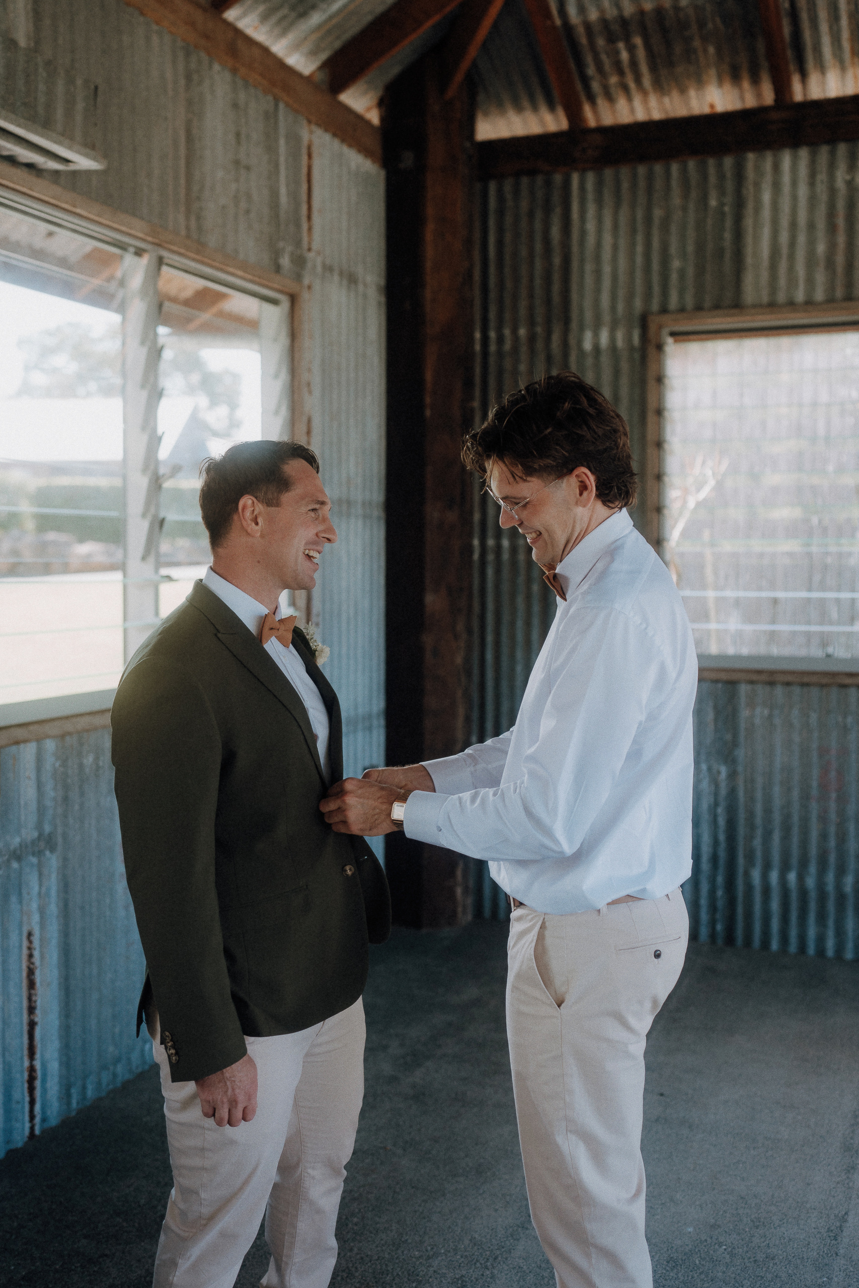 Two men stand in a rustic, corrugated metal room. One man in a white shirt adjusts the jacket of another man wearing a green blazer. Both are smiling.
