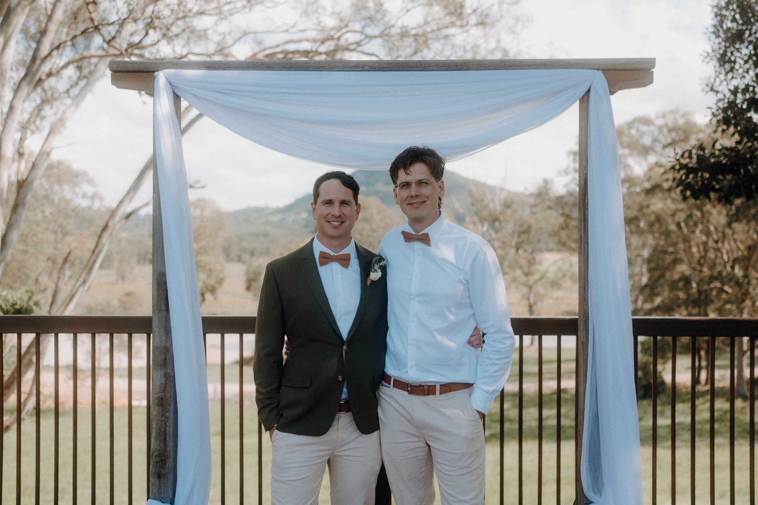 Two men stand side by side under a wedding arch outdoors, both wearing bow ties and light-colored pants, with trees and hills in the background.