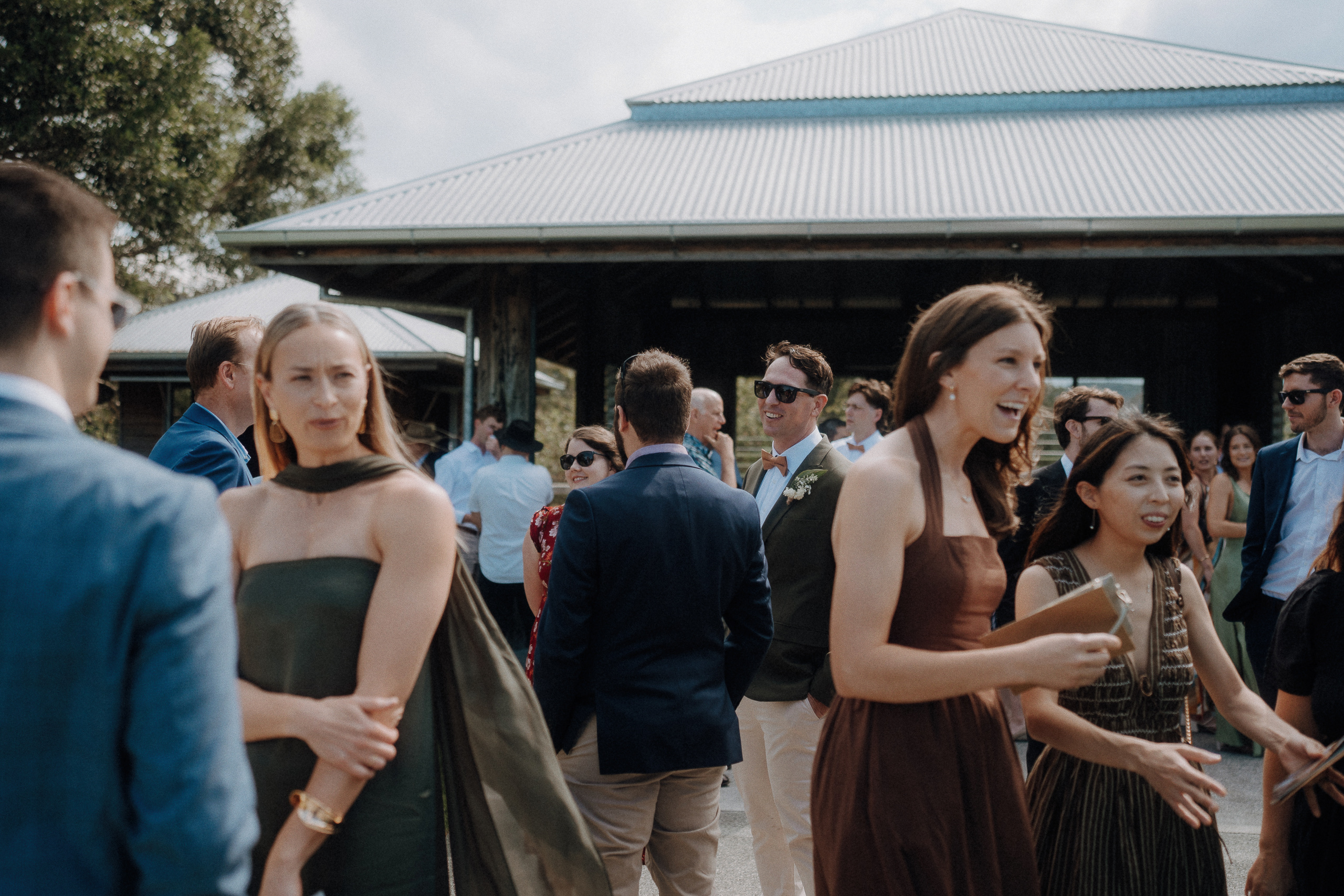 A group of well-dressed people gather and socialize outdoors in front of a building with a metal roof on a sunny day.