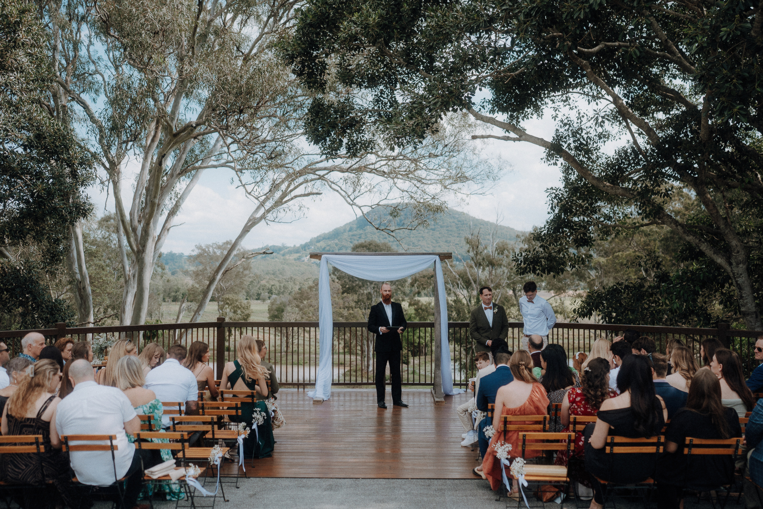 An outdoor wedding ceremony with guests seated facing an officiant standing under a white arch on a wooden deck, surrounded by trees and mountains in the background.