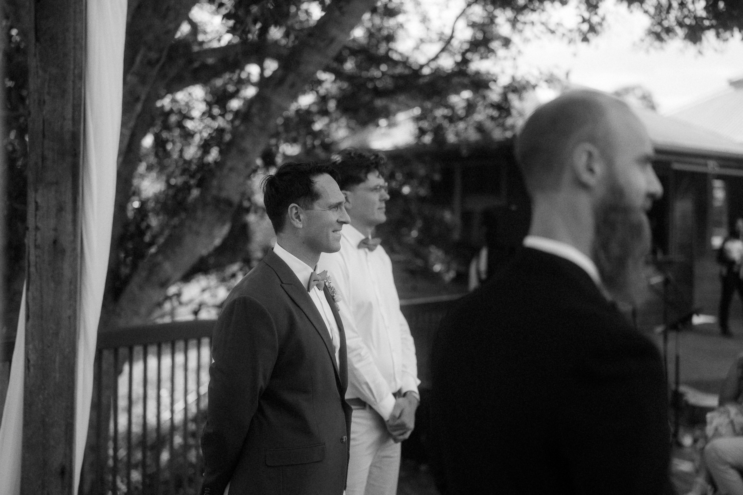 Three men in formal attire stand outdoors near a wooden post and railing, with trees and a building in the background.
