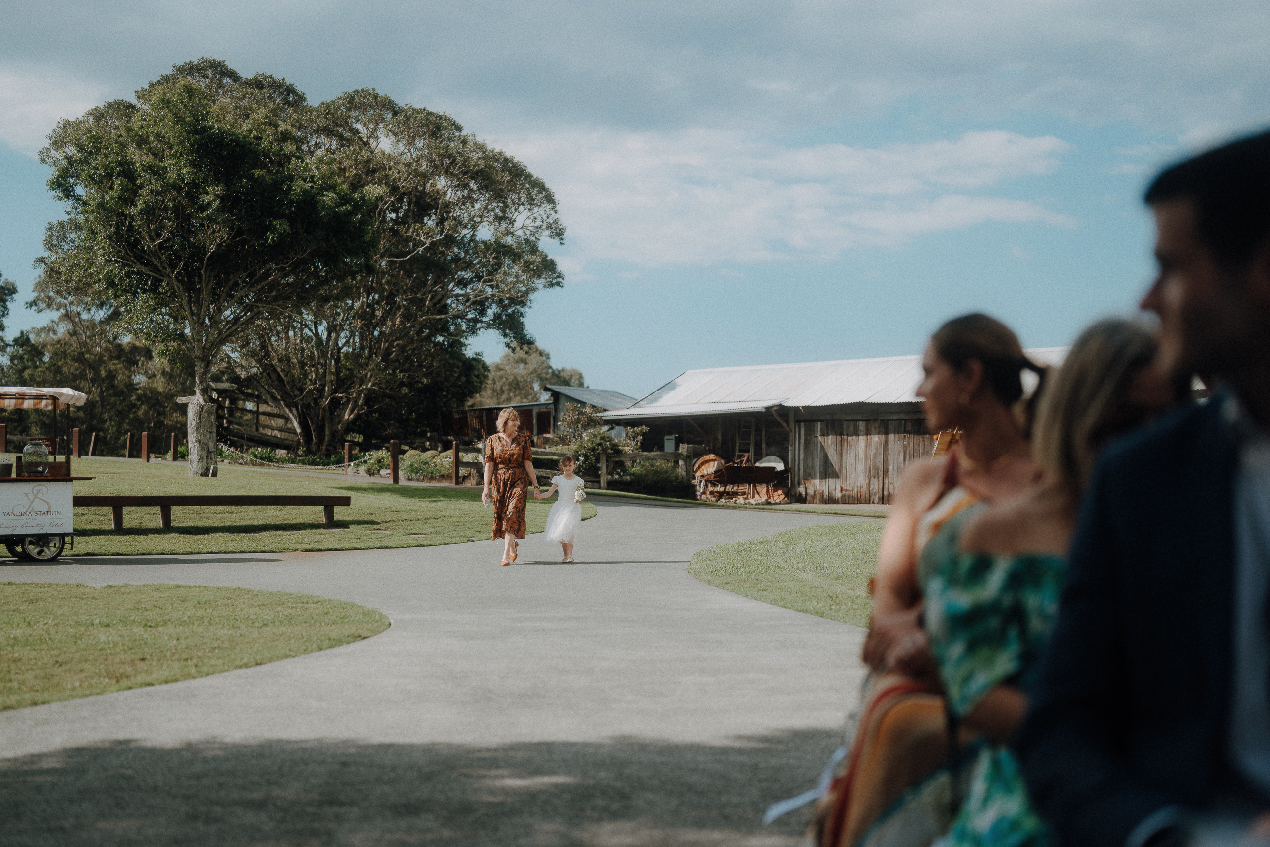 A woman and a child walk down a path toward seated guests at an outdoor event on a sunny day, with trees and rustic buildings in the background.