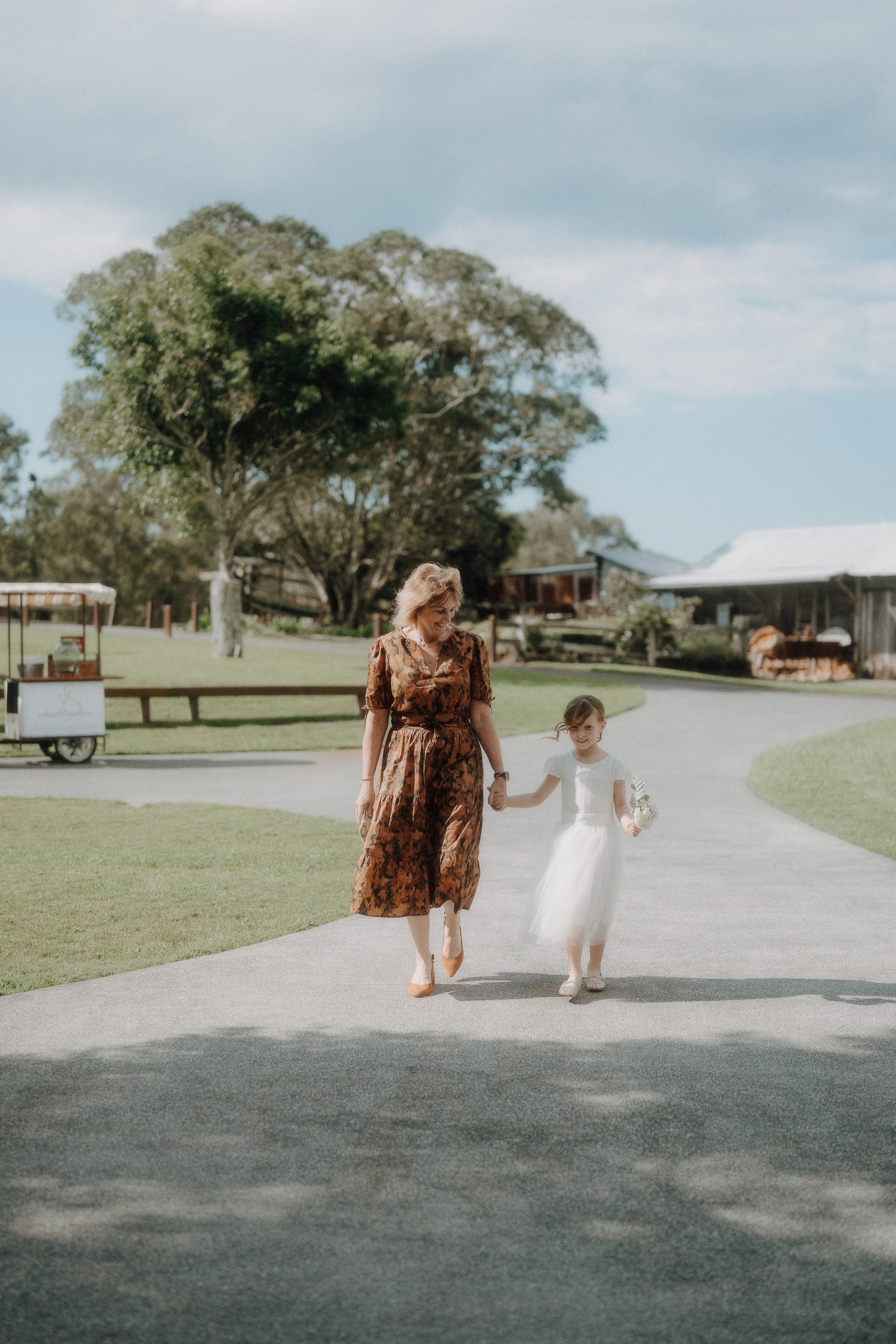A woman in a brown dress walks hand-in-hand with a young girl in a white dress and flower crown on a paved path in a rural outdoor setting.