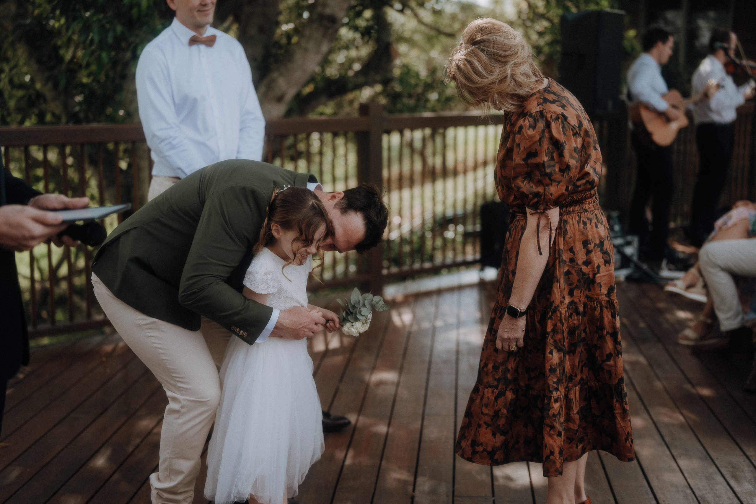 A man bends down to hug a young girl in a white dress while an older woman in a patterned dress stands nearby on a wooden deck during an outdoor event.