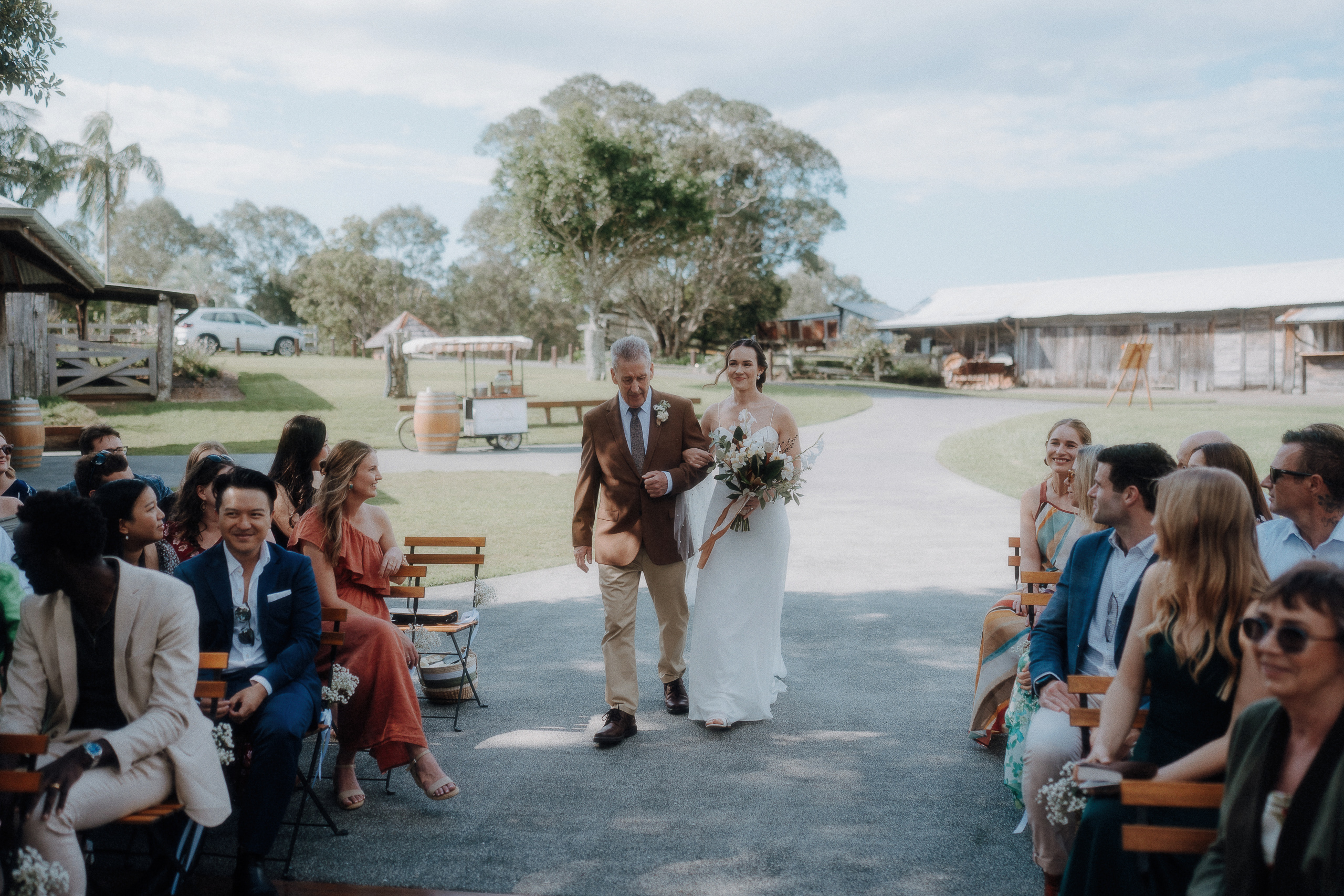 A bride in a white dress walks down an outdoor aisle with an older man, surrounded by seated guests at a rustic wedding venue.