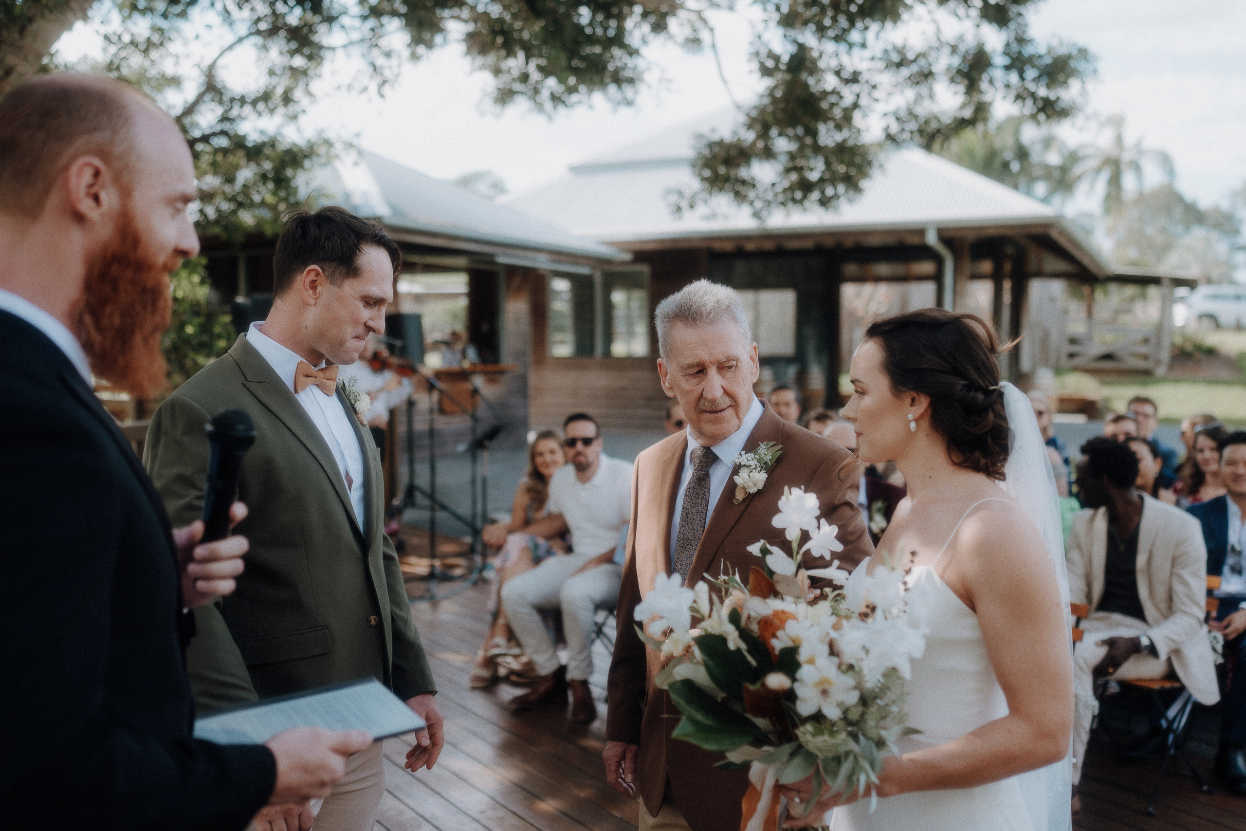 A bride holding a bouquet stands next to an older man, facing a groom and officiant, during an outdoor wedding ceremony with guests seated in the background.
