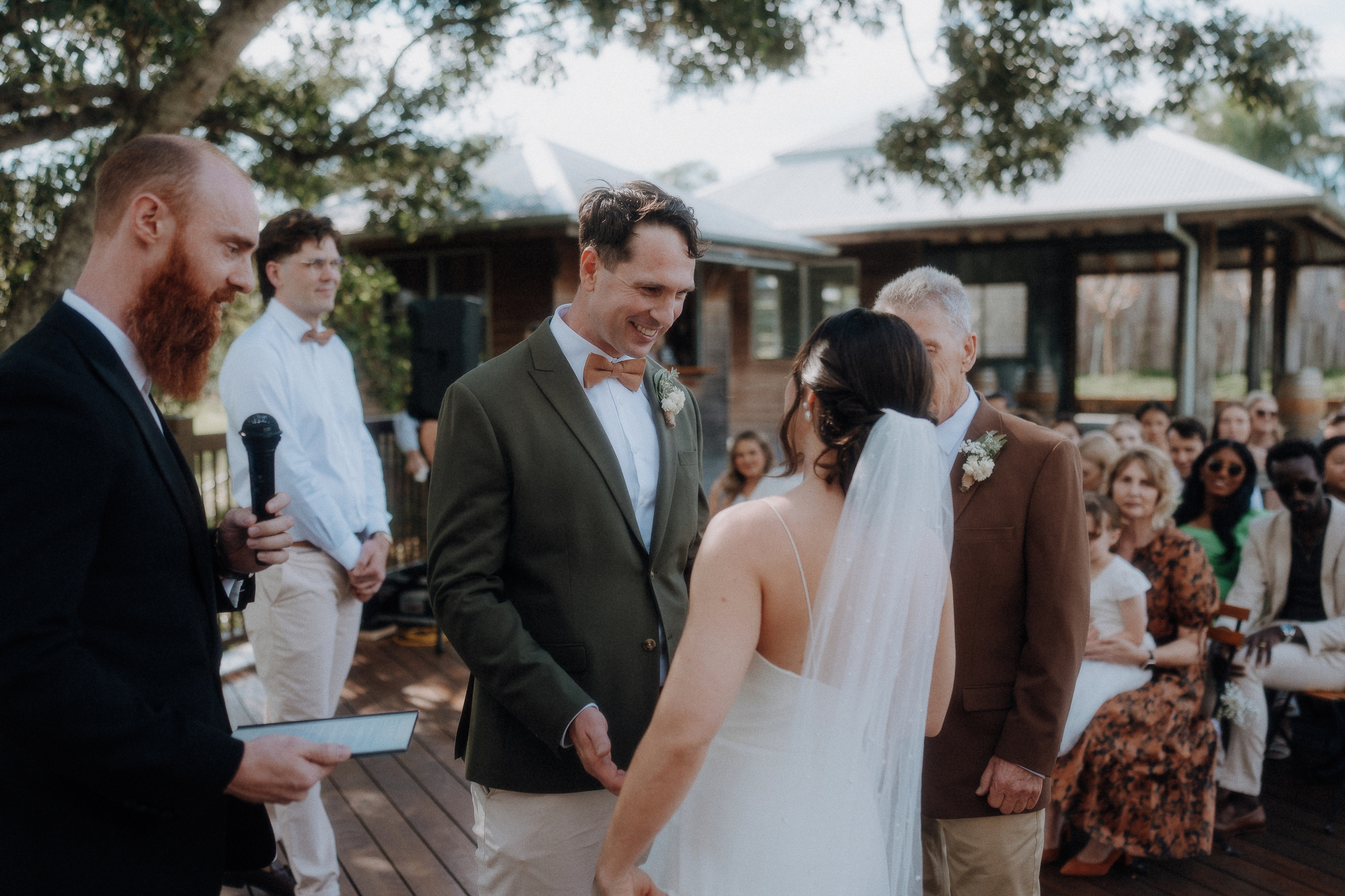 A bride and groom stand facing each other during an outdoor wedding ceremony, with a man holding a microphone and guests seated in the background.