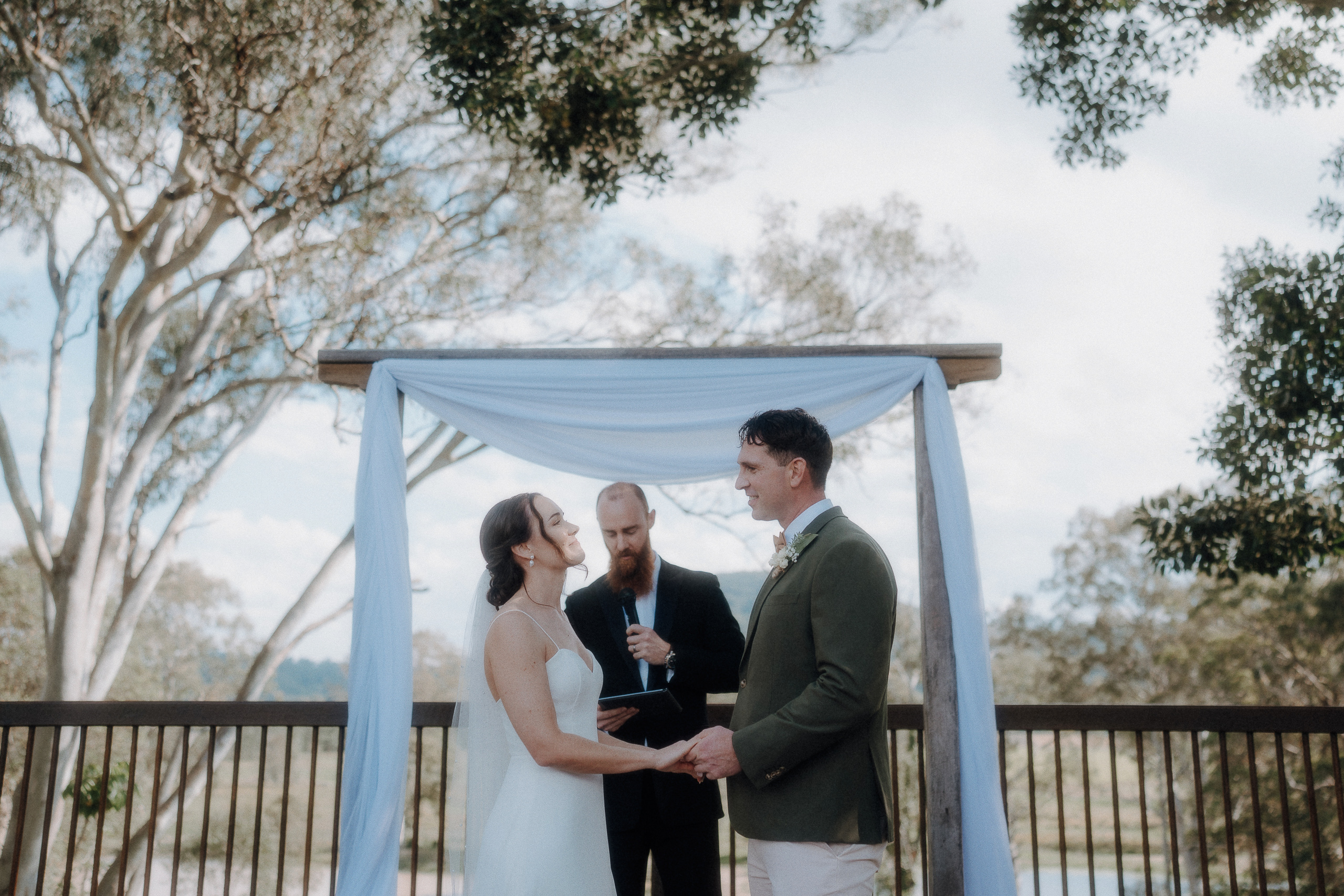 A bride and groom stand facing each other and hold hands under a white canopy, with an officiant standing behind them outdoors.