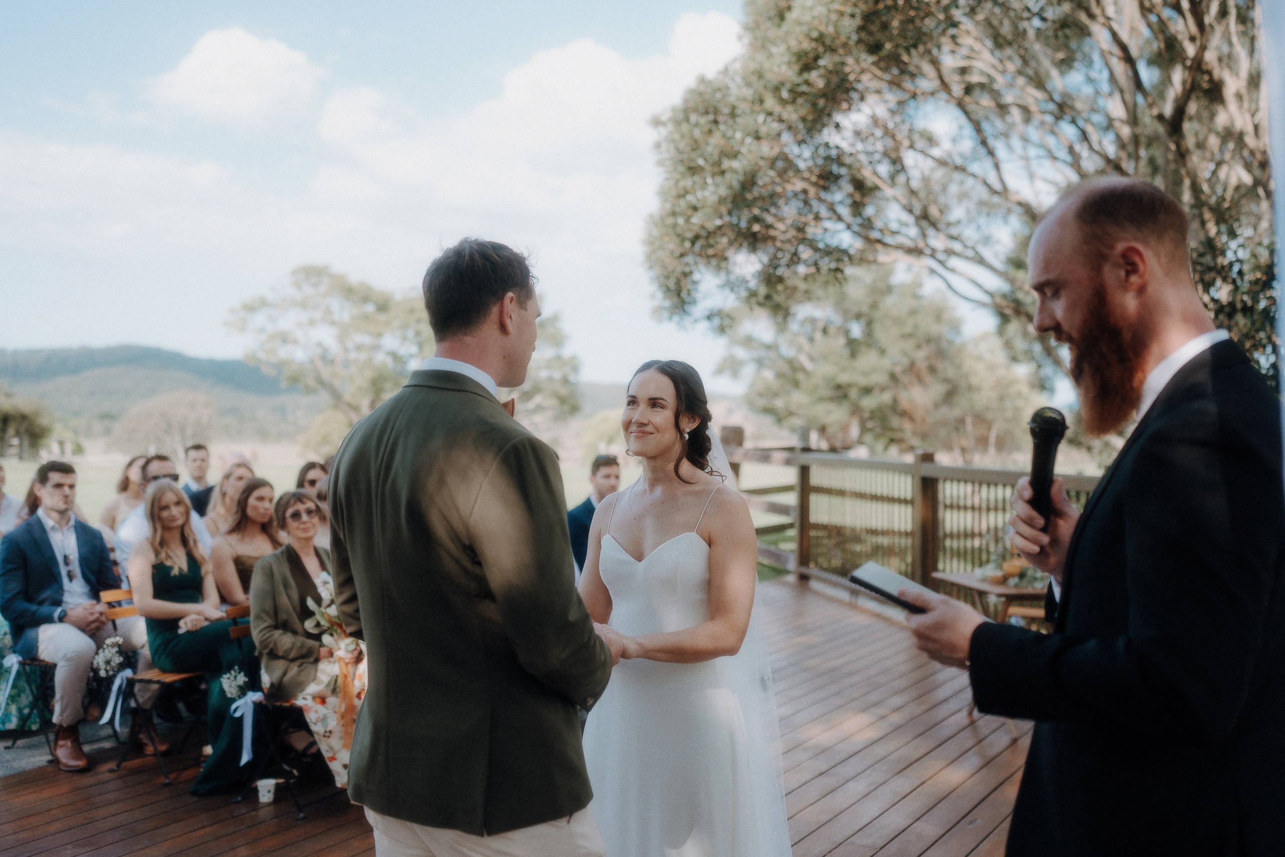 A bride and groom stand facing each other during an outdoor wedding ceremony as a man reads from a microphone, with guests seated in the background.