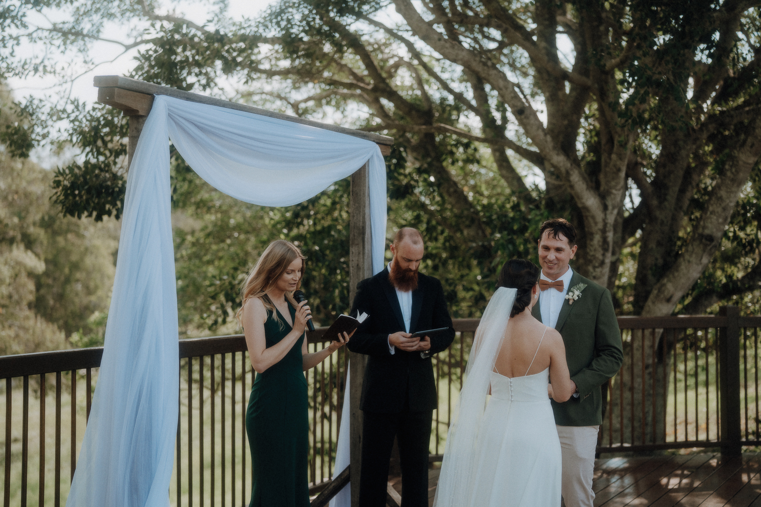A bride and groom stand under a draped arch outdoors, facing each other, with a man officiating and a woman reading from a book nearby. Trees and greenery are visible in the background.