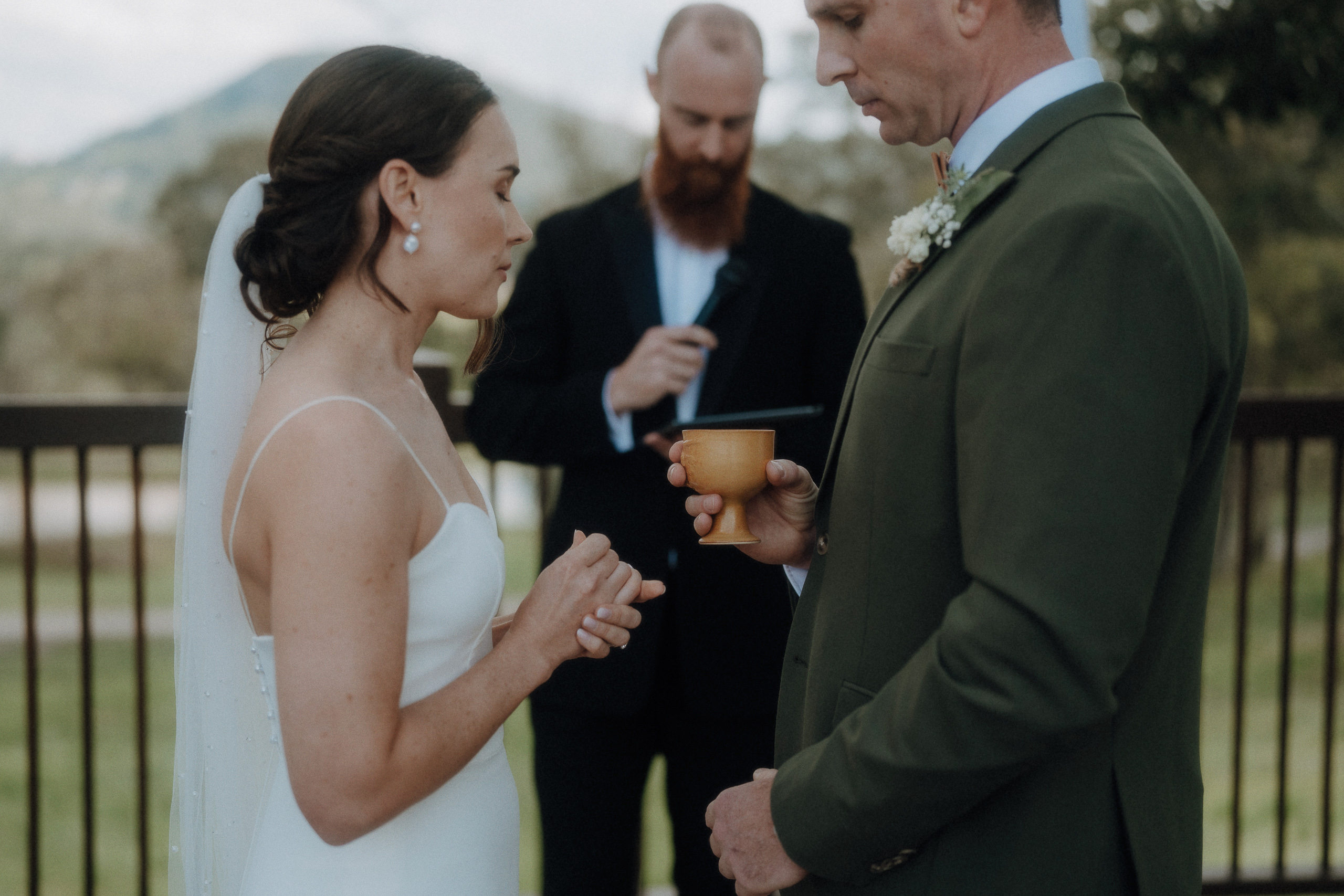 A bride and groom stand facing each other holding a cup, with an officiant in the background, during an outdoor wedding ceremony.