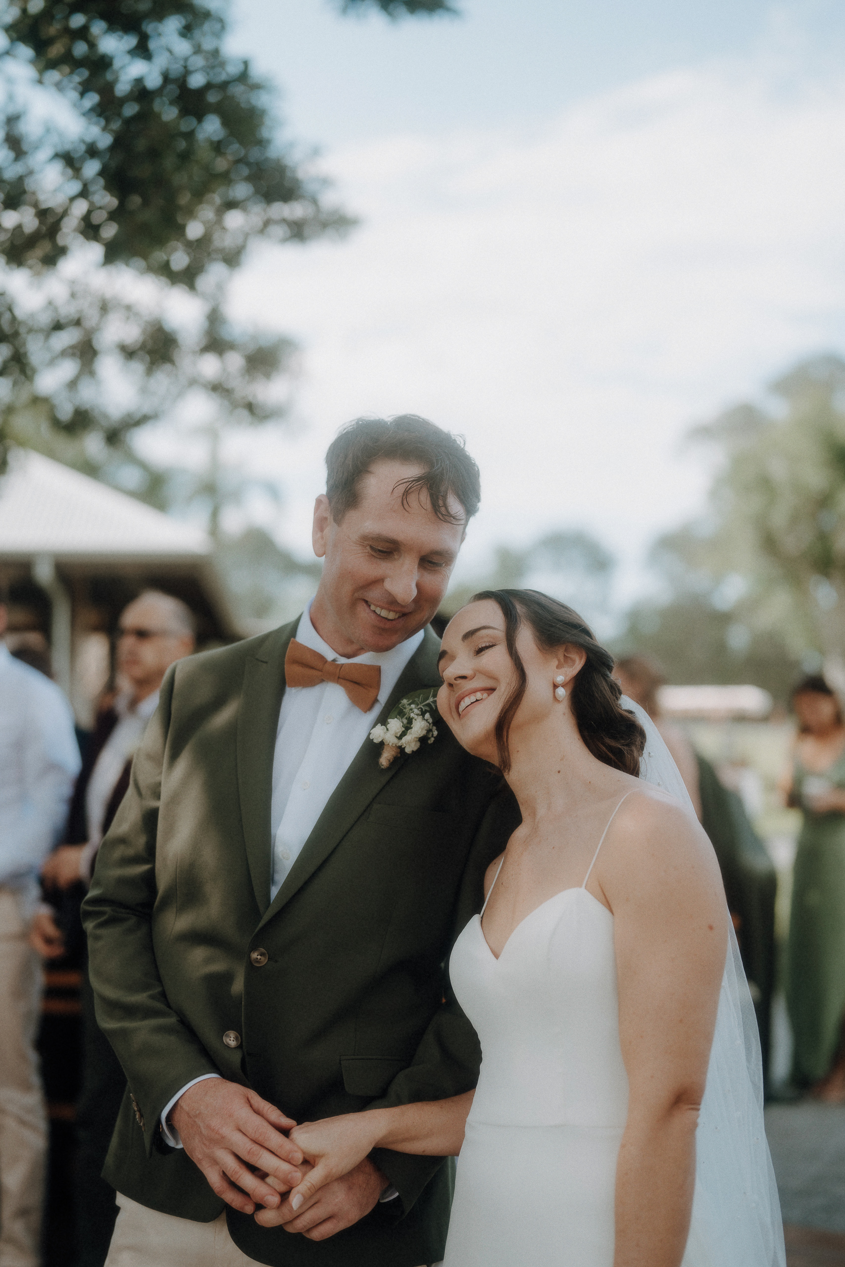 A bride in a white dress leans her head on the groom’s shoulder; they are smiling and holding hands outdoors with guests in the background.