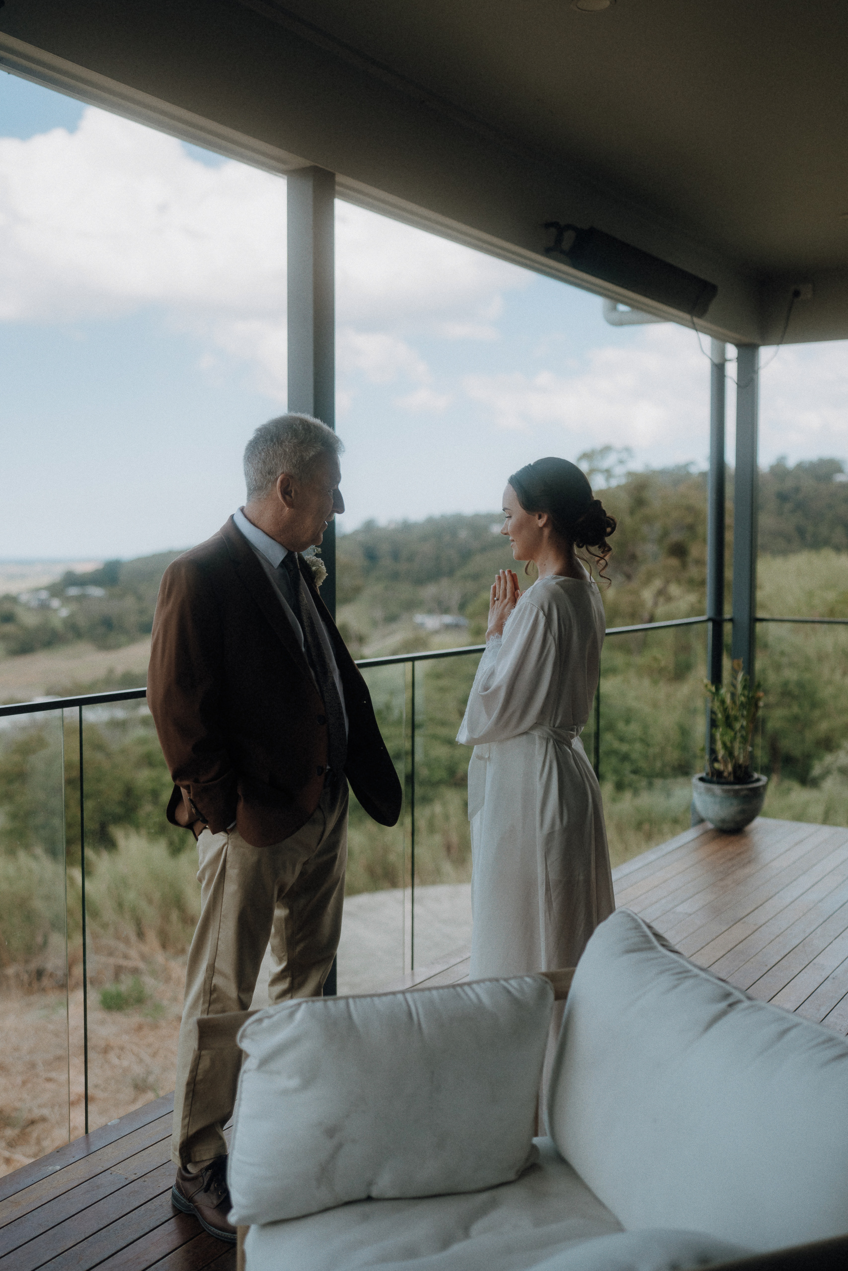 A man in a suit and a woman in a white robe stand on a balcony, facing each other, with a scenic landscape visible in the background.
