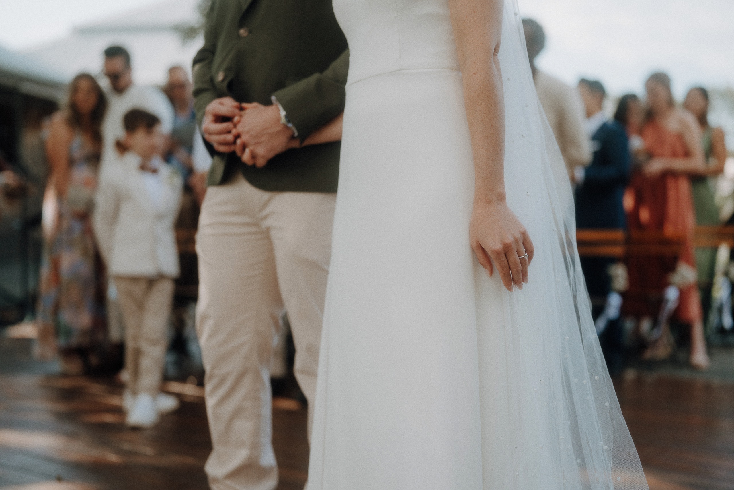 Two people stand side by side, one in a white dress with a veil and the other in light-colored pants and a green jacket, at an outdoor event with guests in the background.