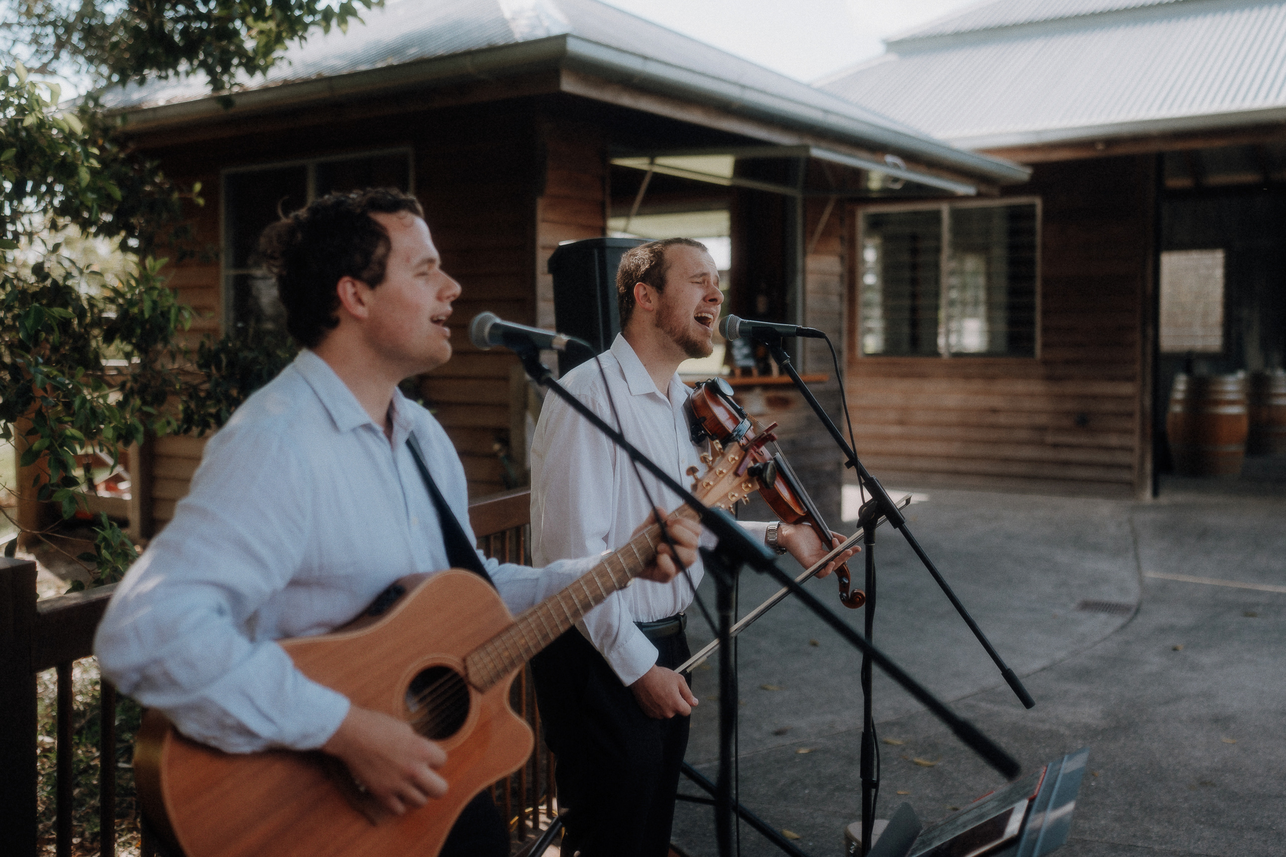 Two men perform outdoors; one plays acoustic guitar and sings, the other plays violin and sings, both standing in front of microphones near a wooden building.