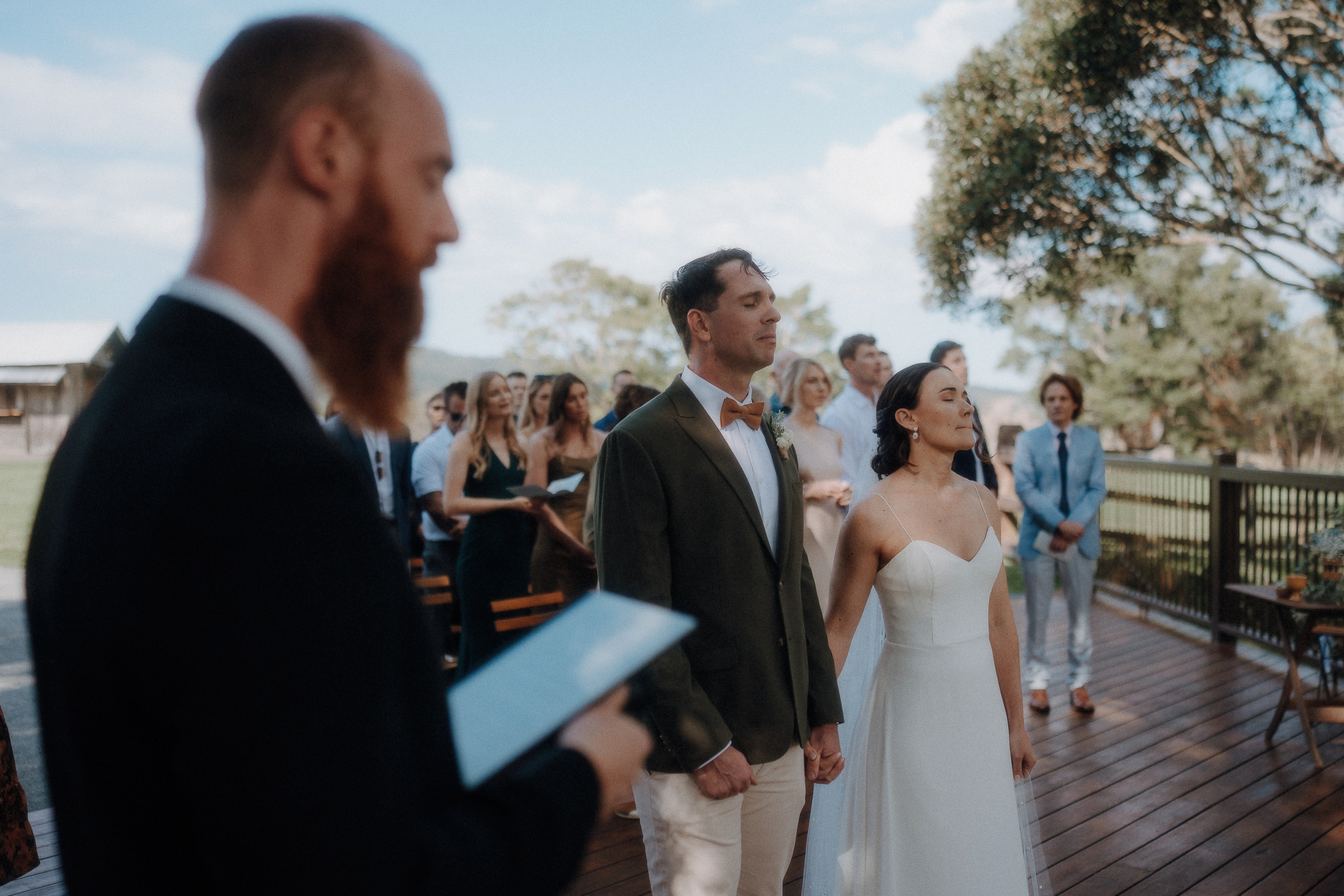 A couple stands holding hands during an outdoor wedding ceremony, with an officiant reading from a book and guests watching in the background.