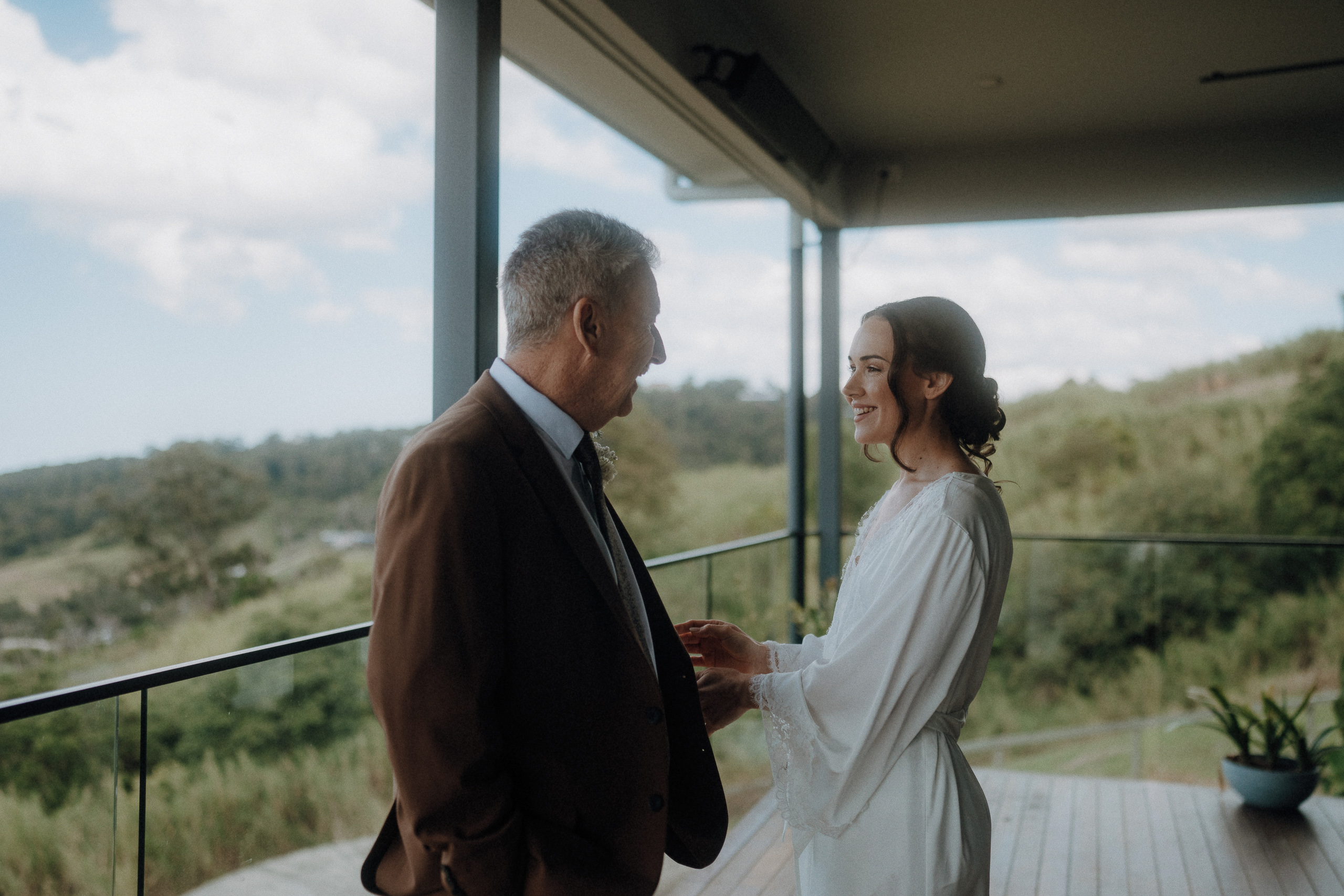 A man in a suit and a woman in a white dress stand facing each other and smiling on a covered outdoor deck with a scenic landscape in the background.