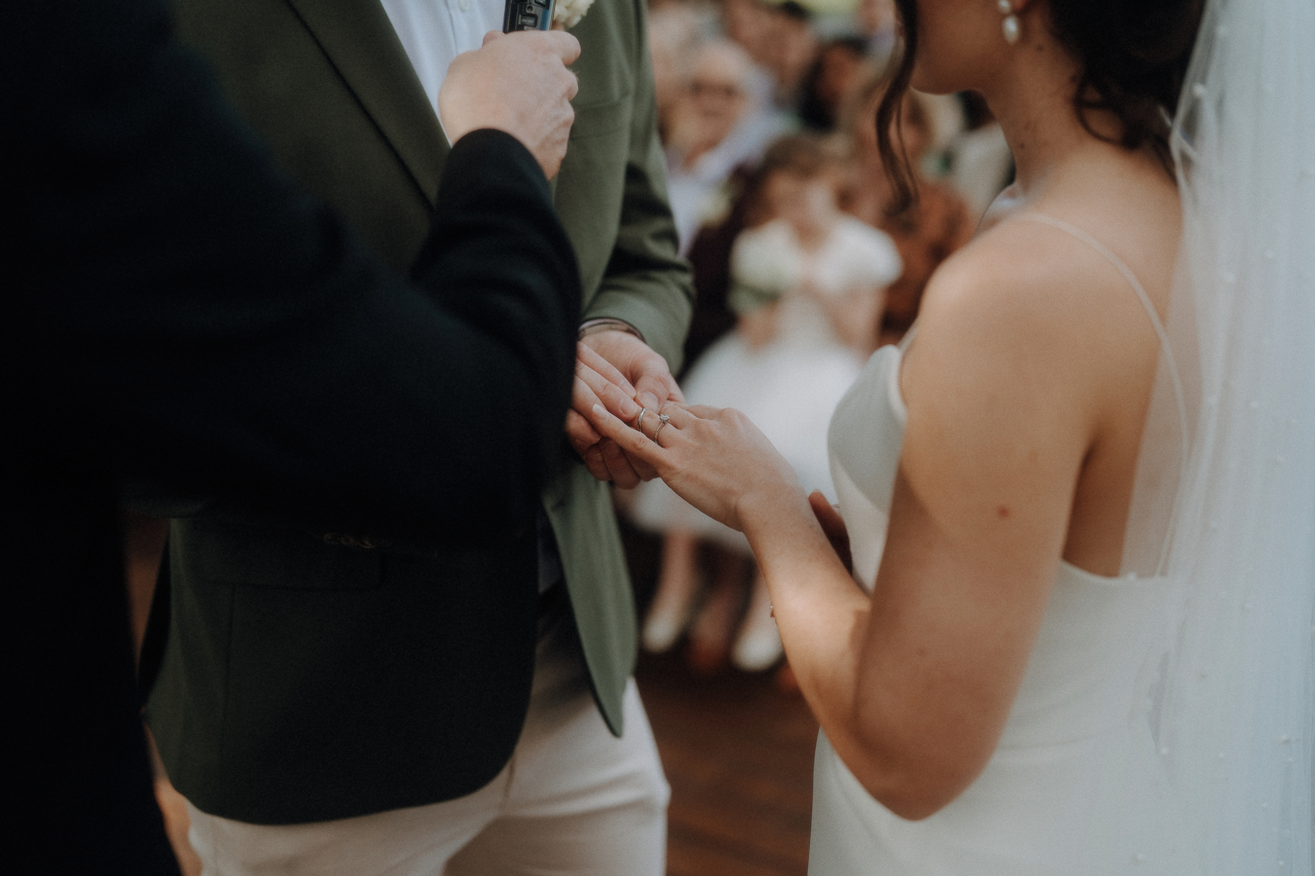 A bride and groom exchange rings during a wedding ceremony, with the officiant holding a microphone and guests blurred in the background.