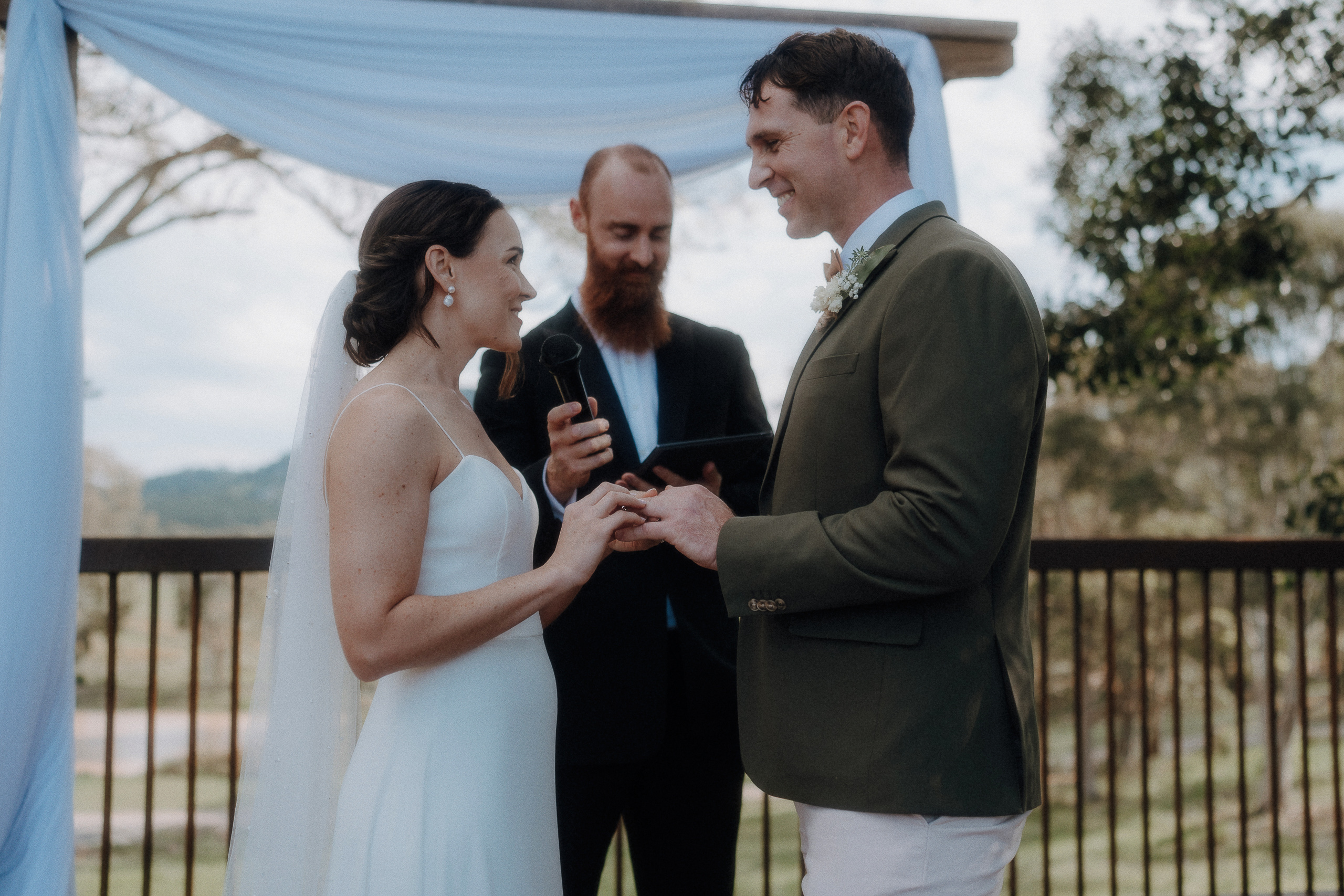 A bride and groom exchange rings during an outdoor wedding ceremony, with an officiant standing behind them holding a microphone and script.