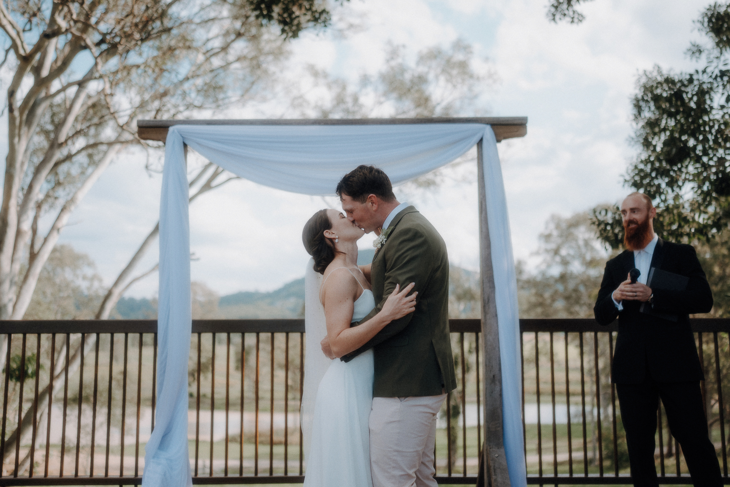 A bride and groom share a kiss under an outdoor wedding arch while a man in a suit stands nearby holding a microphone.