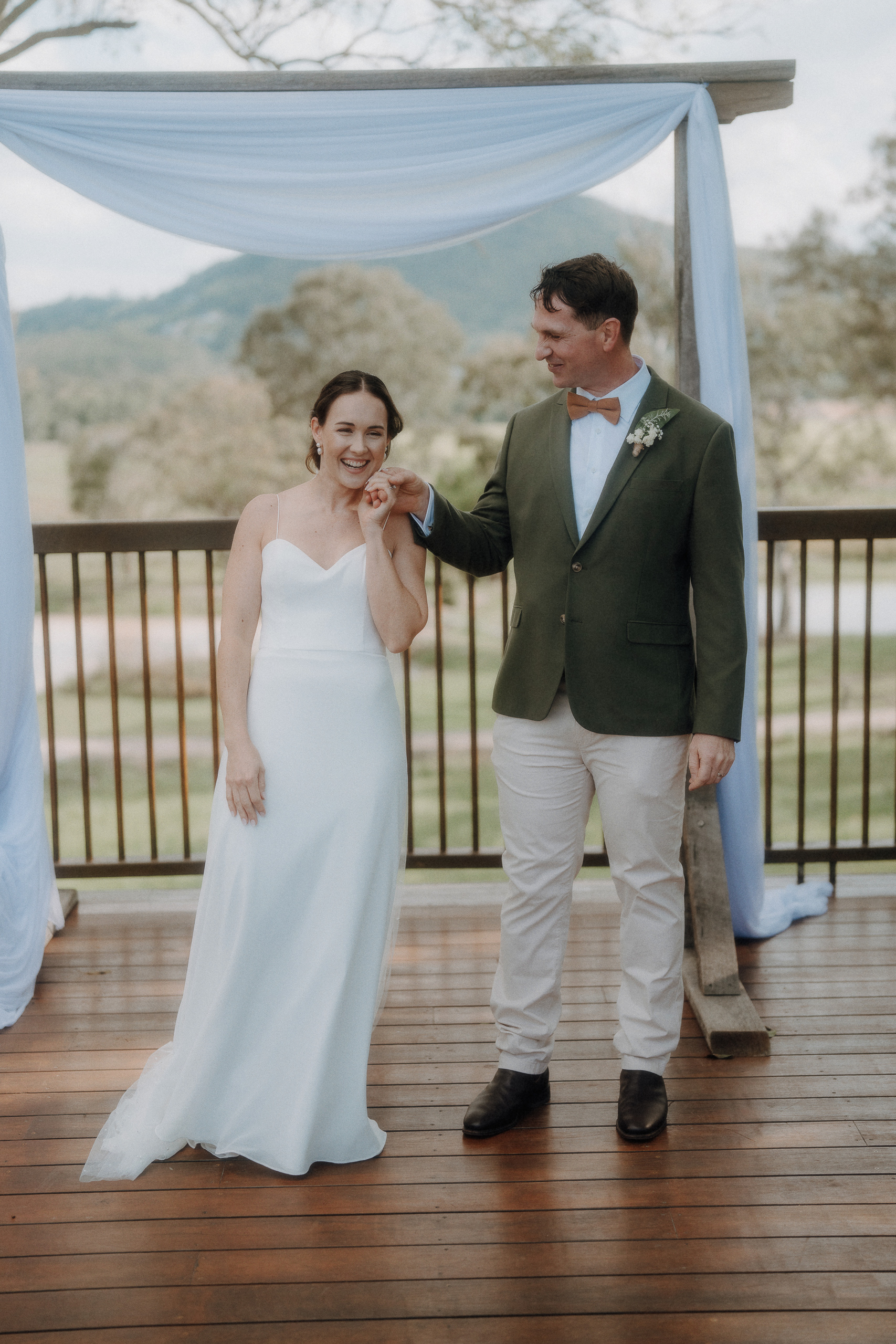A bride in a white dress and a groom in a green jacket stand under an outdoor arch, smiling and holding hands on a wooden deck.