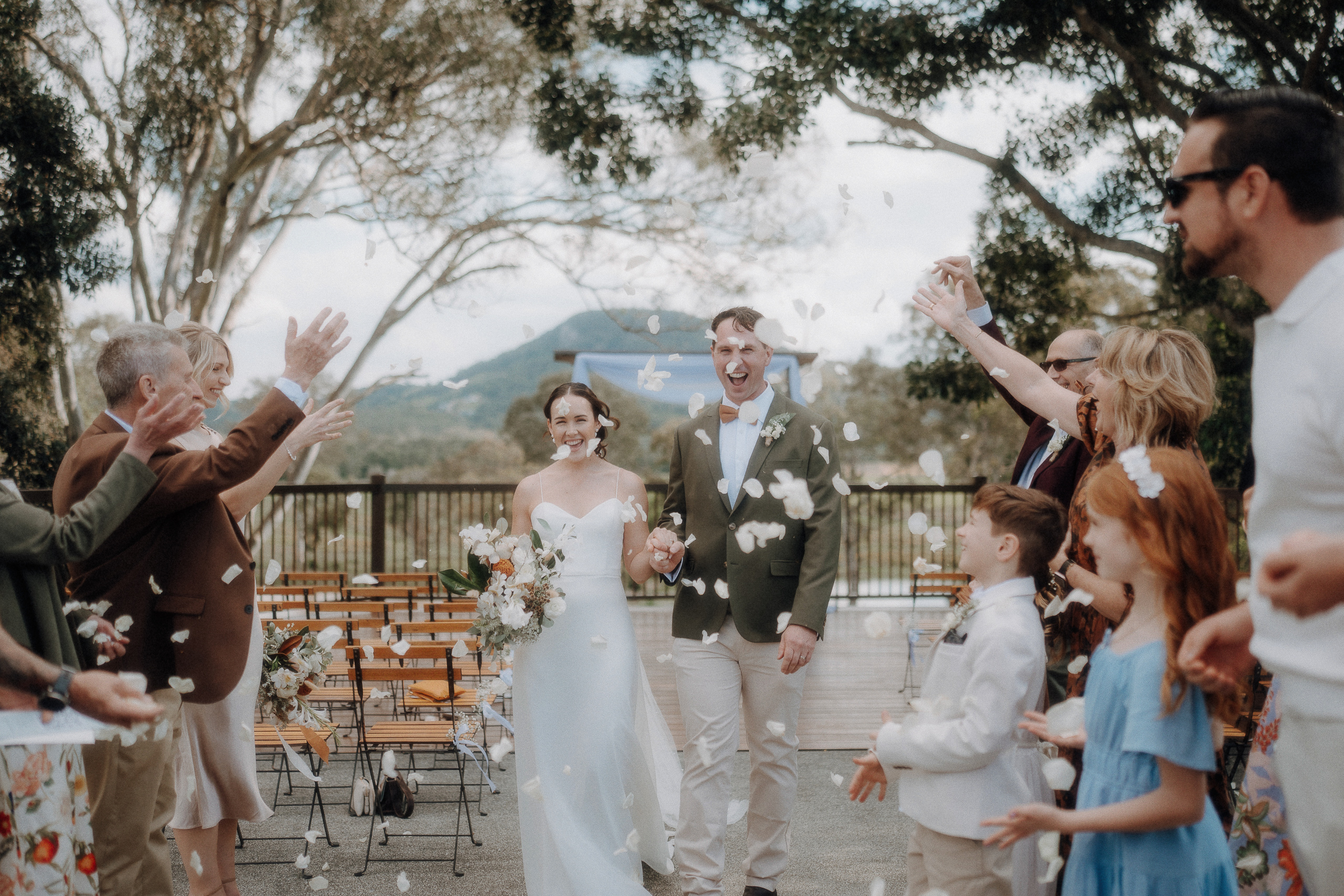 A newlywed couple walks down the aisle outdoors as guests throw flower petals and celebrate.