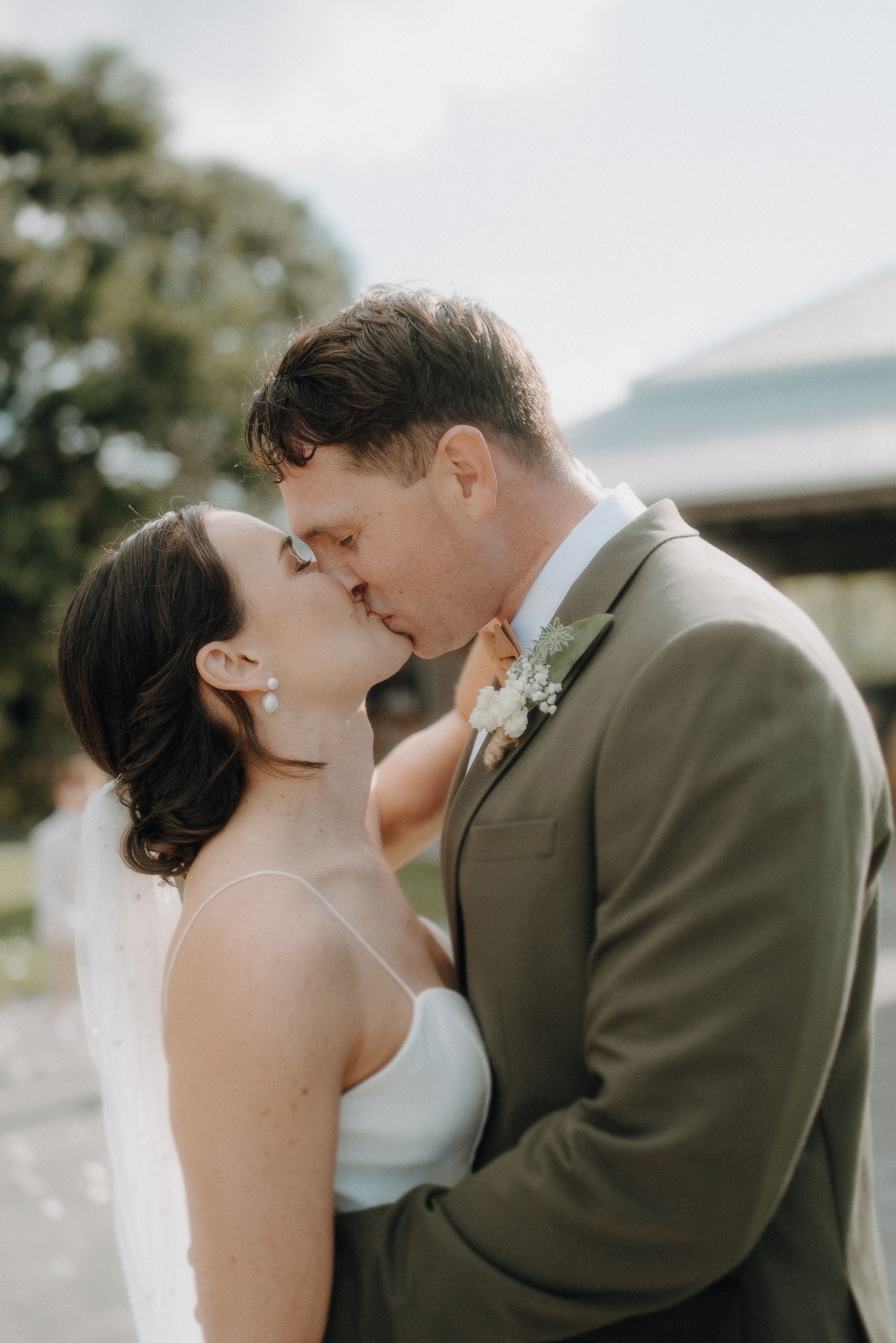 A bride and groom share a kiss outdoors, dressed in wedding attire, with greenery and a building in the blurred background.