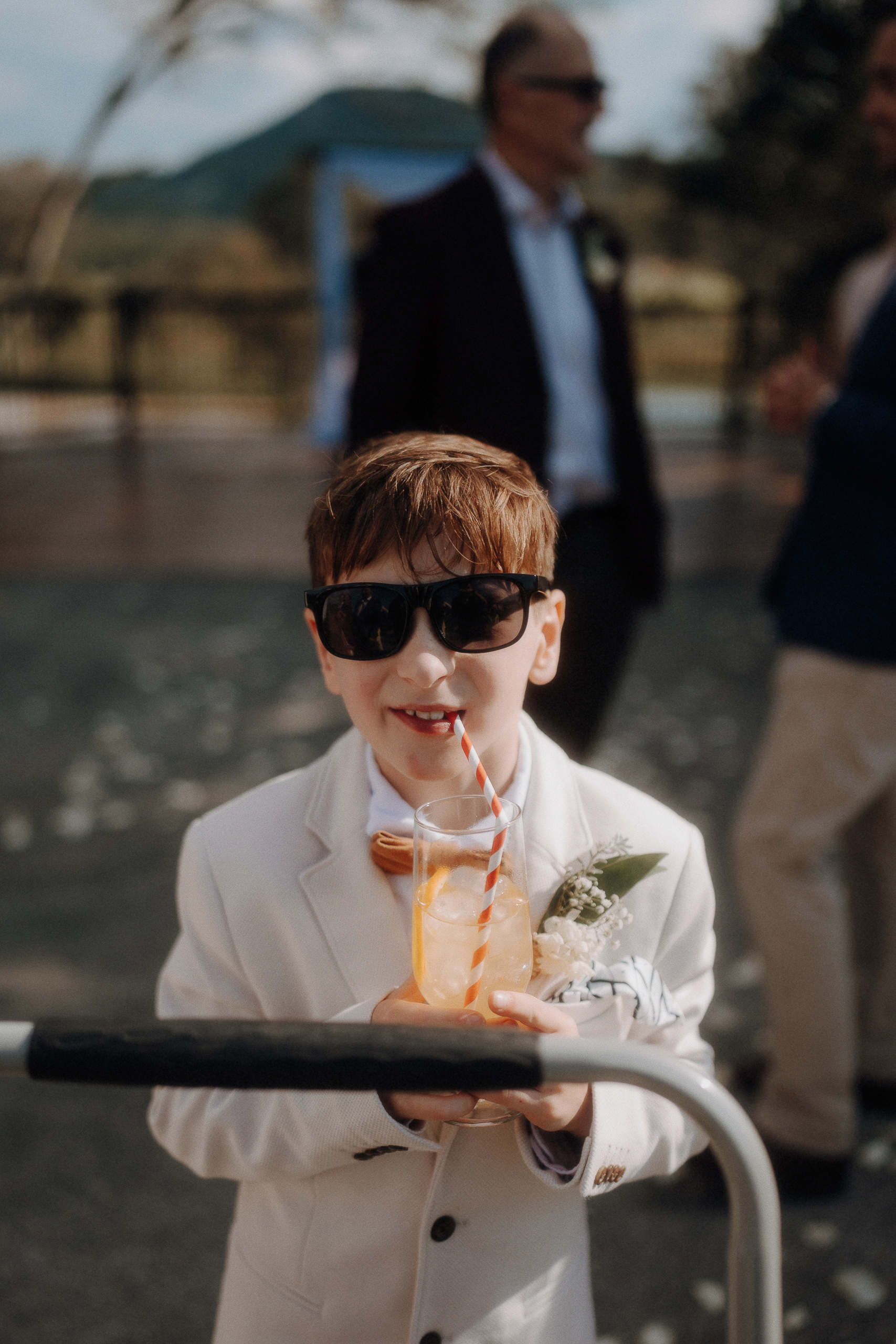 A young boy in a white suit and sunglasses drinks from a glass with a striped straw at an outdoor event.
