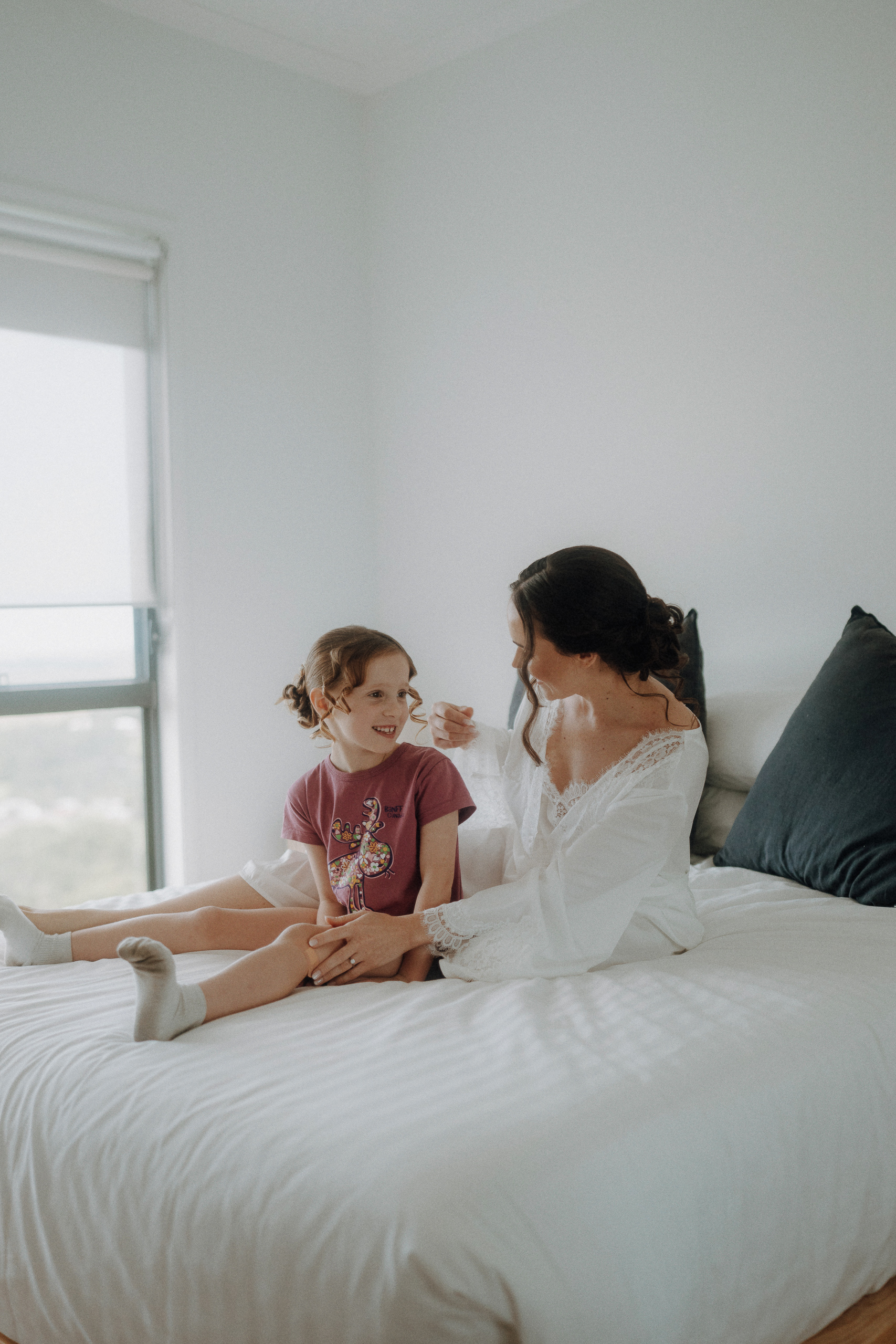 A woman in a white robe sits on a bed next to a young girl in a t-shirt and socks. They are smiling at each other in a bright, softly lit room.