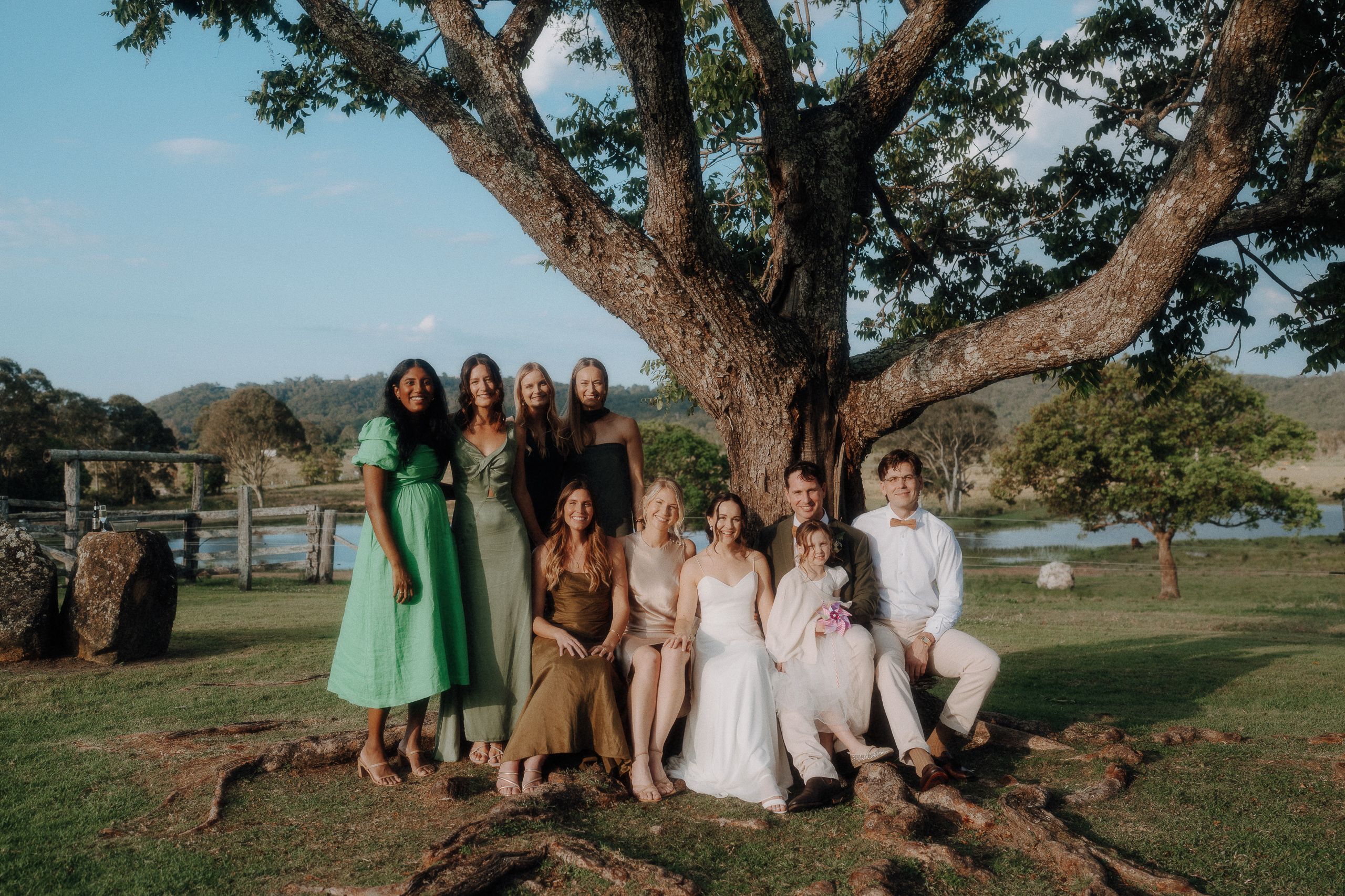 A group of ten people, dressed in light and formal attire, pose together under a large tree outdoors with hills and a pond in the background.