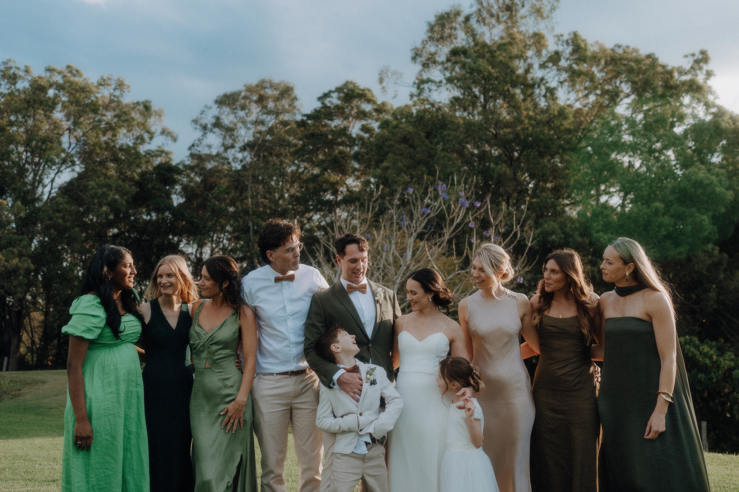 A wedding party poses outdoors, with the bride and groom in the center, surrounded by adults and children dressed in formal attire, against a backdrop of trees.