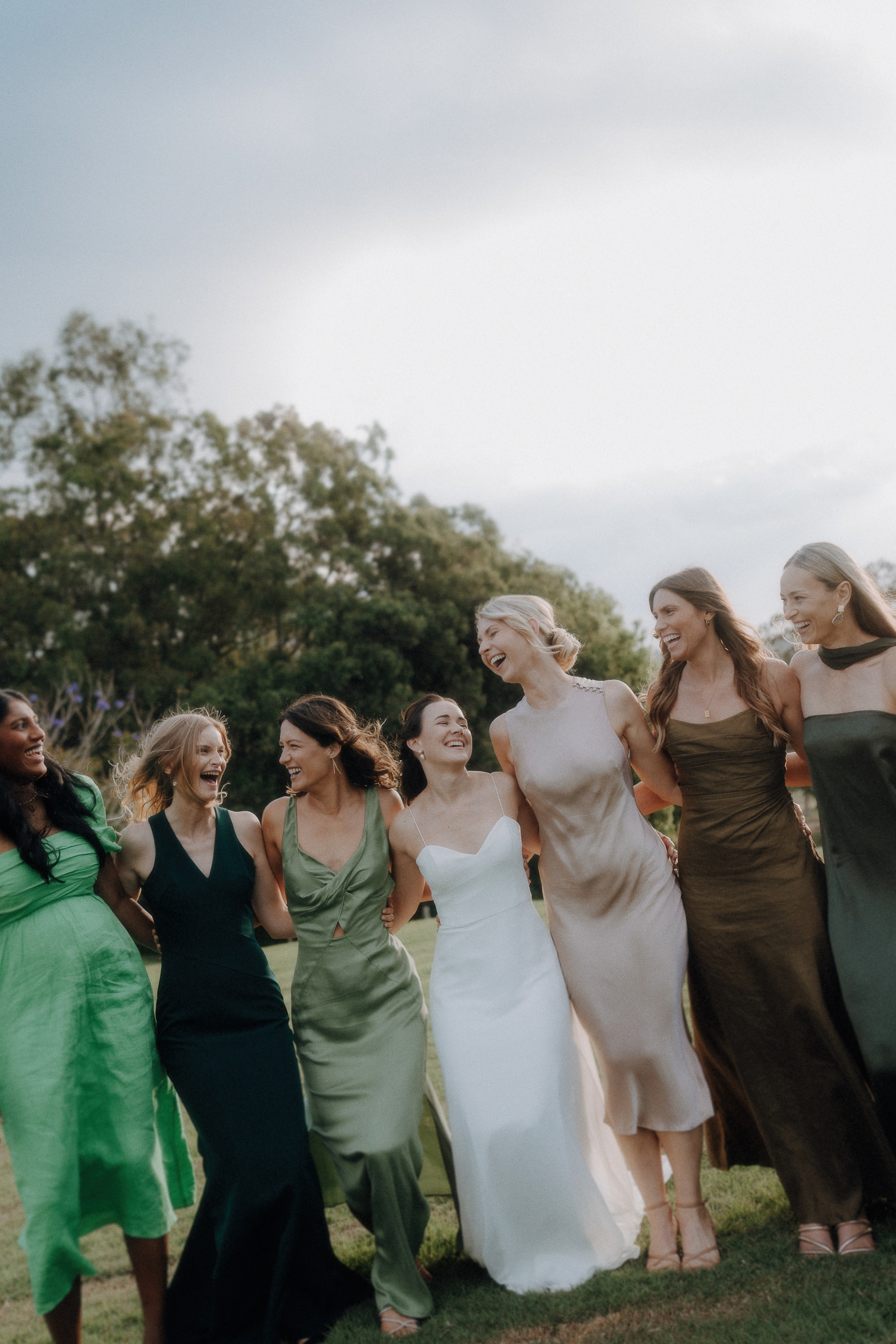 A group of women in dresses, including a bride in white, stand together on grass outdoors, smiling and linking arms. Trees and sky are visible in the background.