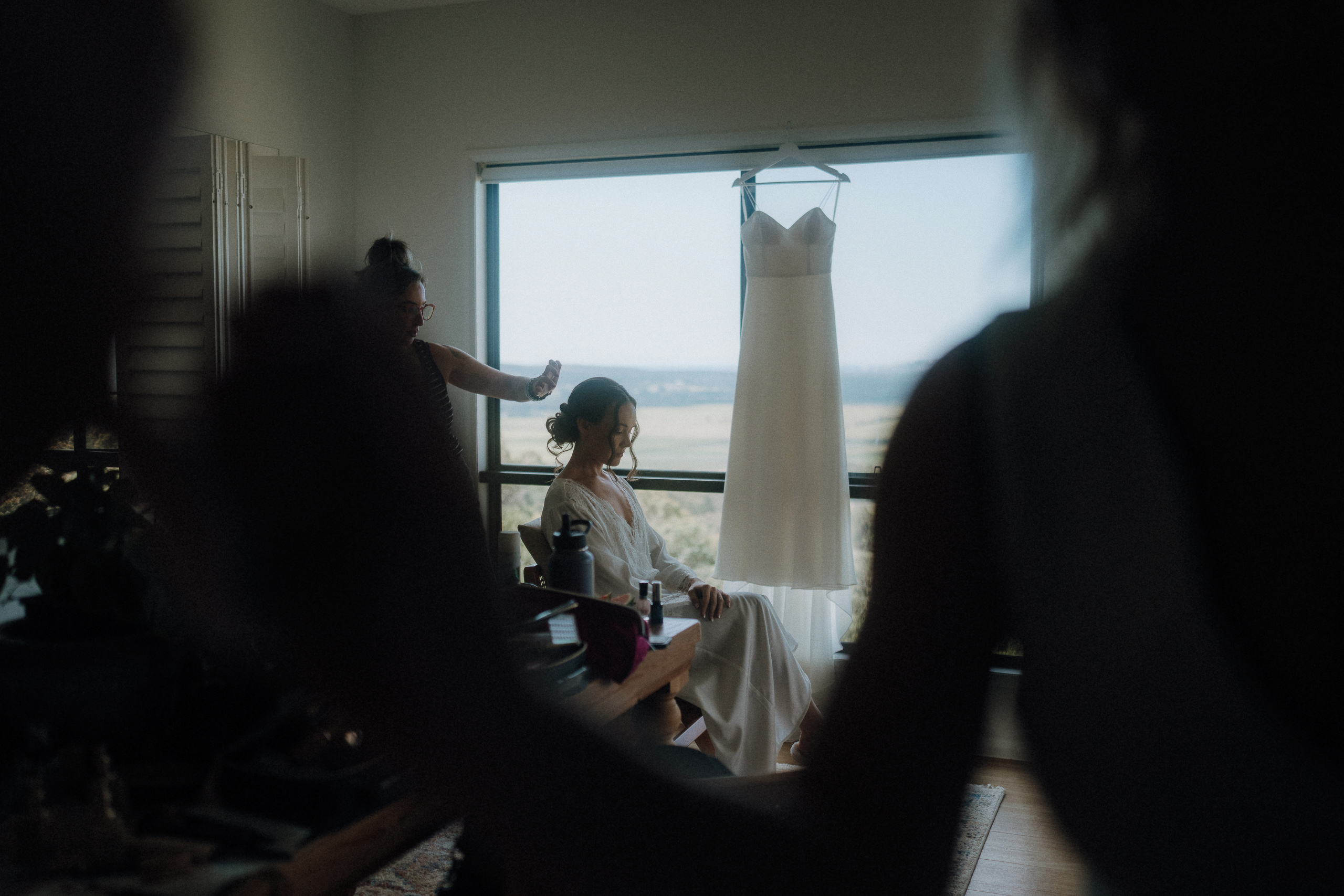 A woman sits in front of a window while another person styles her hair; a white dress hangs nearby.