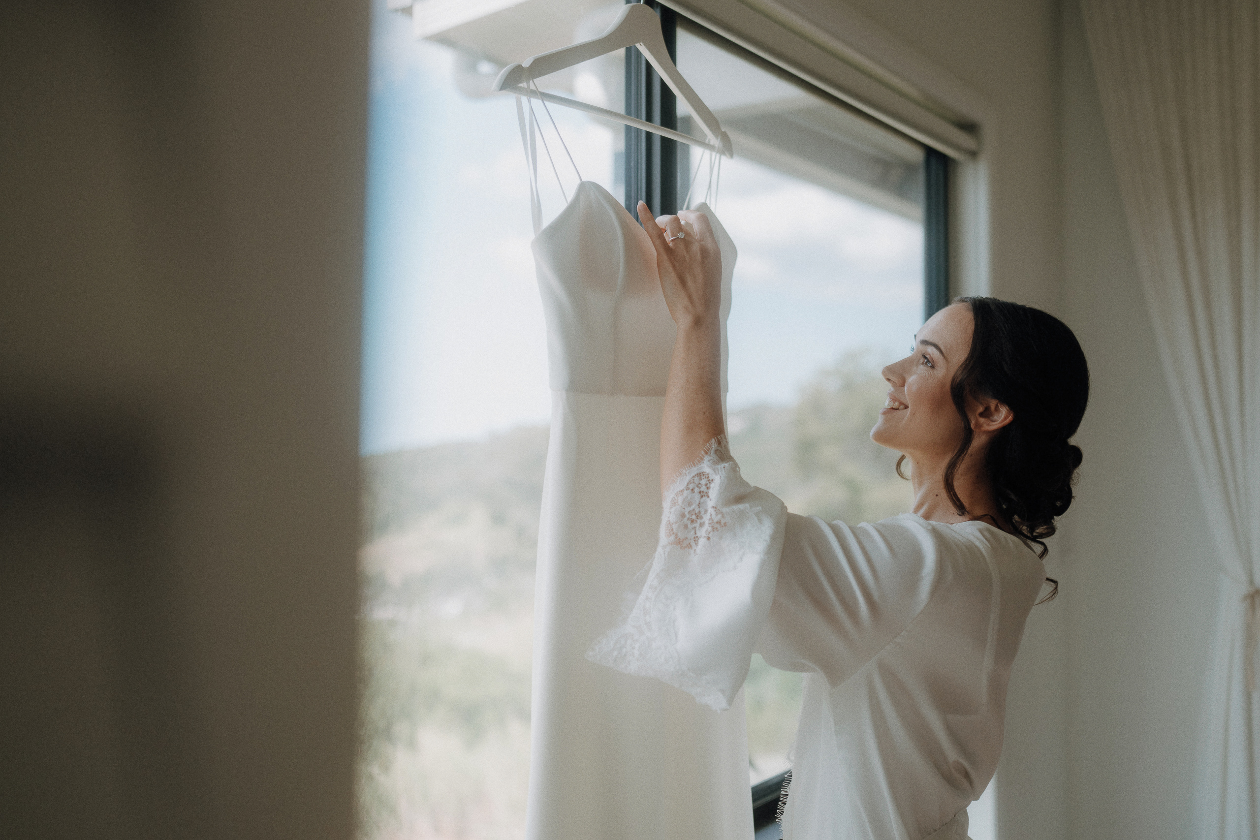 A woman in a white robe stands by a window, smiling as she holds a white dress hanging on a hanger.