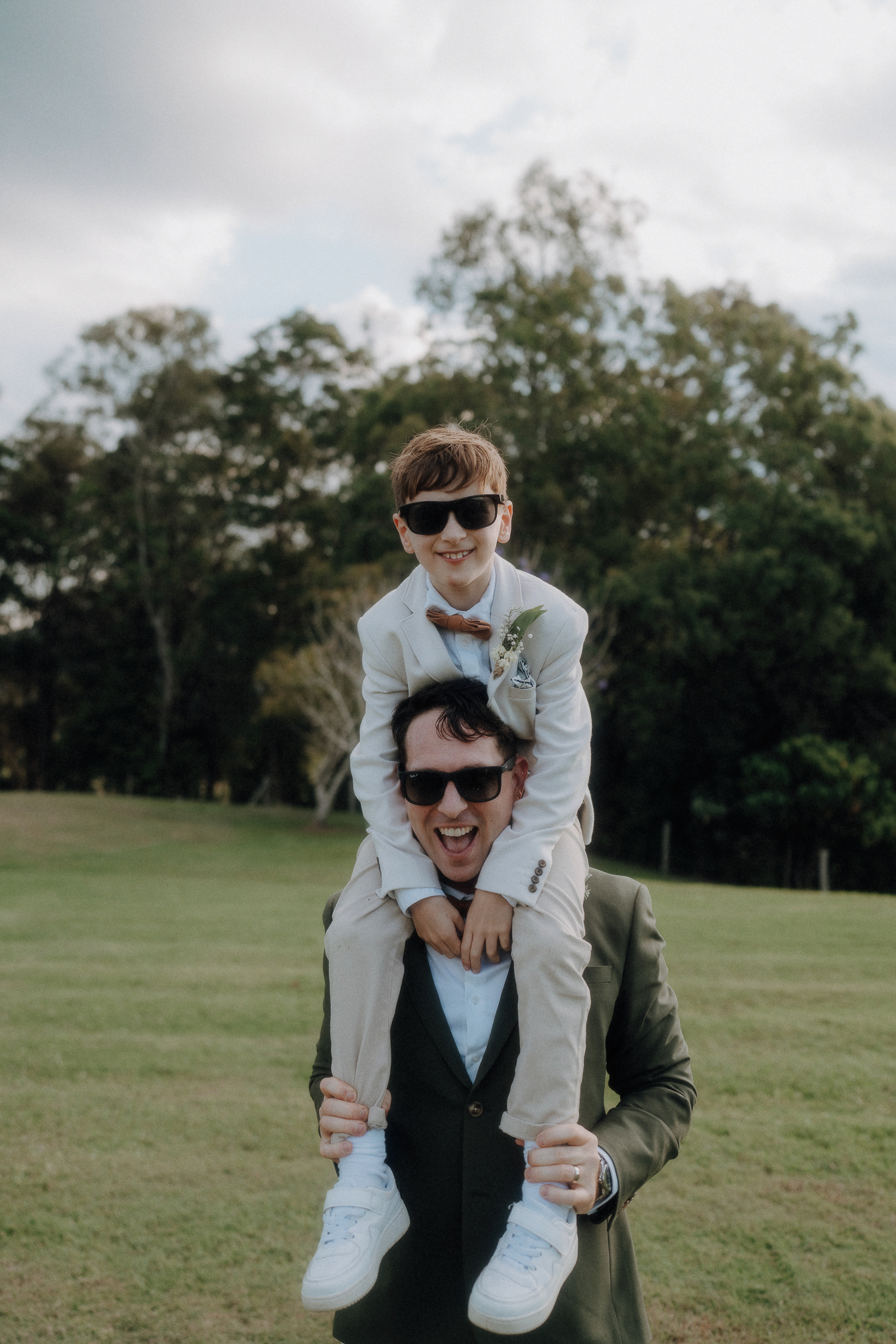 A man in a suit carries a young boy in a white suit and sunglasses on his shoulders in an open grassy area with trees in the background.