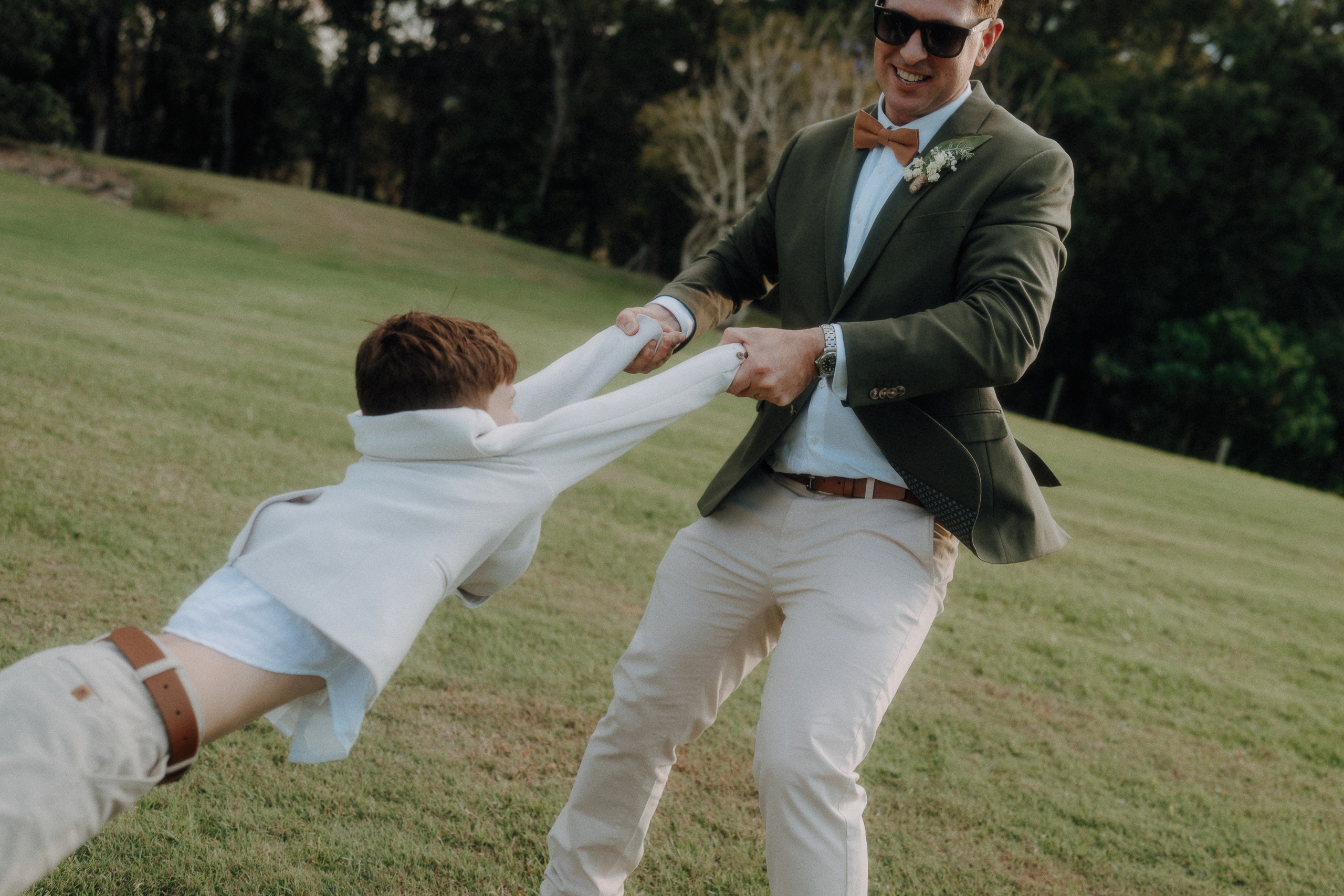 An adult in a suit and sunglasses swings a smiling child by the arms in a grassy outdoor area.