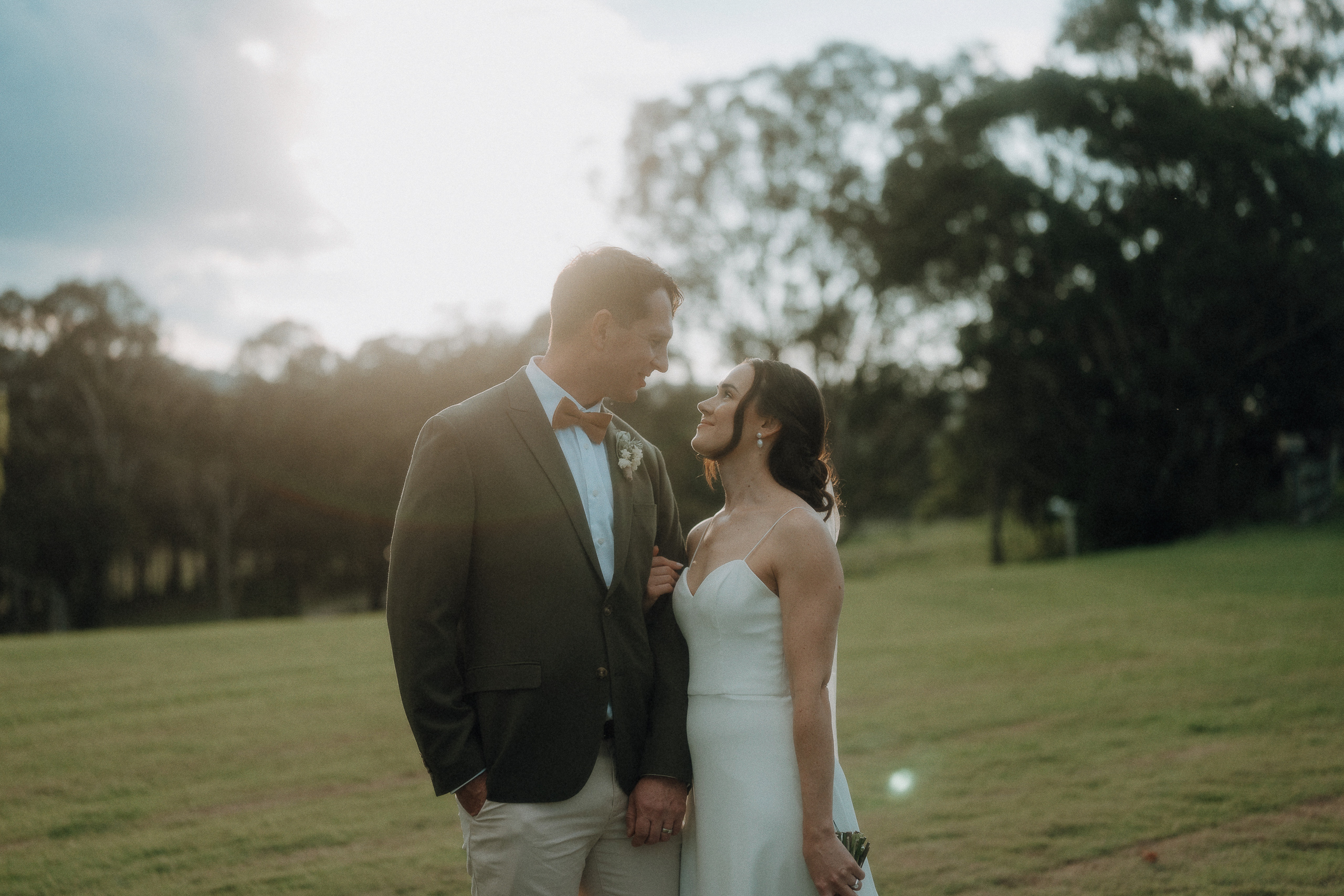 A couple dressed in wedding attire stands outdoors on a grassy field, looking at each other with trees and sunlight in the background.