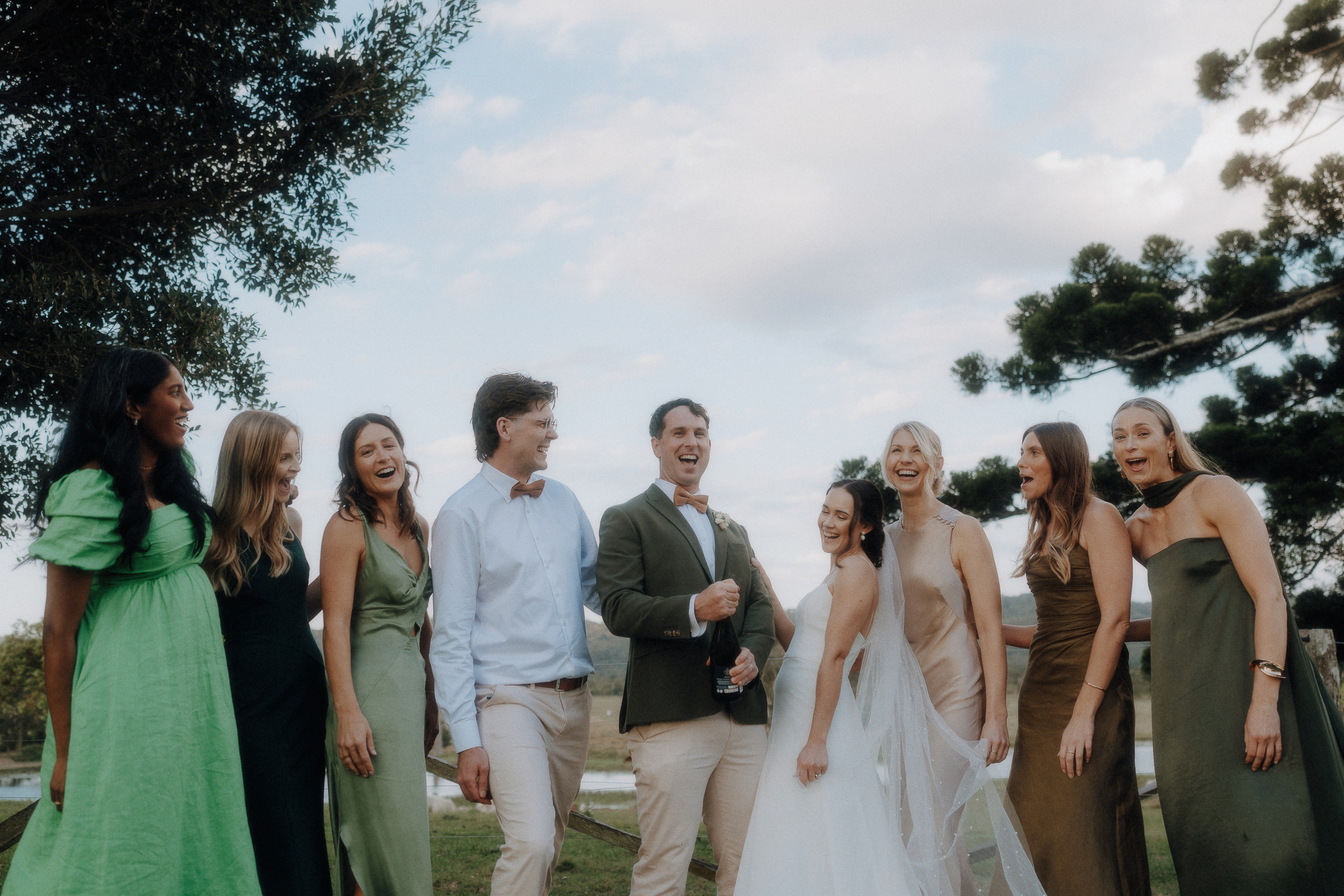 A group of people, dressed in formal and semi-formal attire, stand outdoors smiling and laughing, with trees and sky in the background.