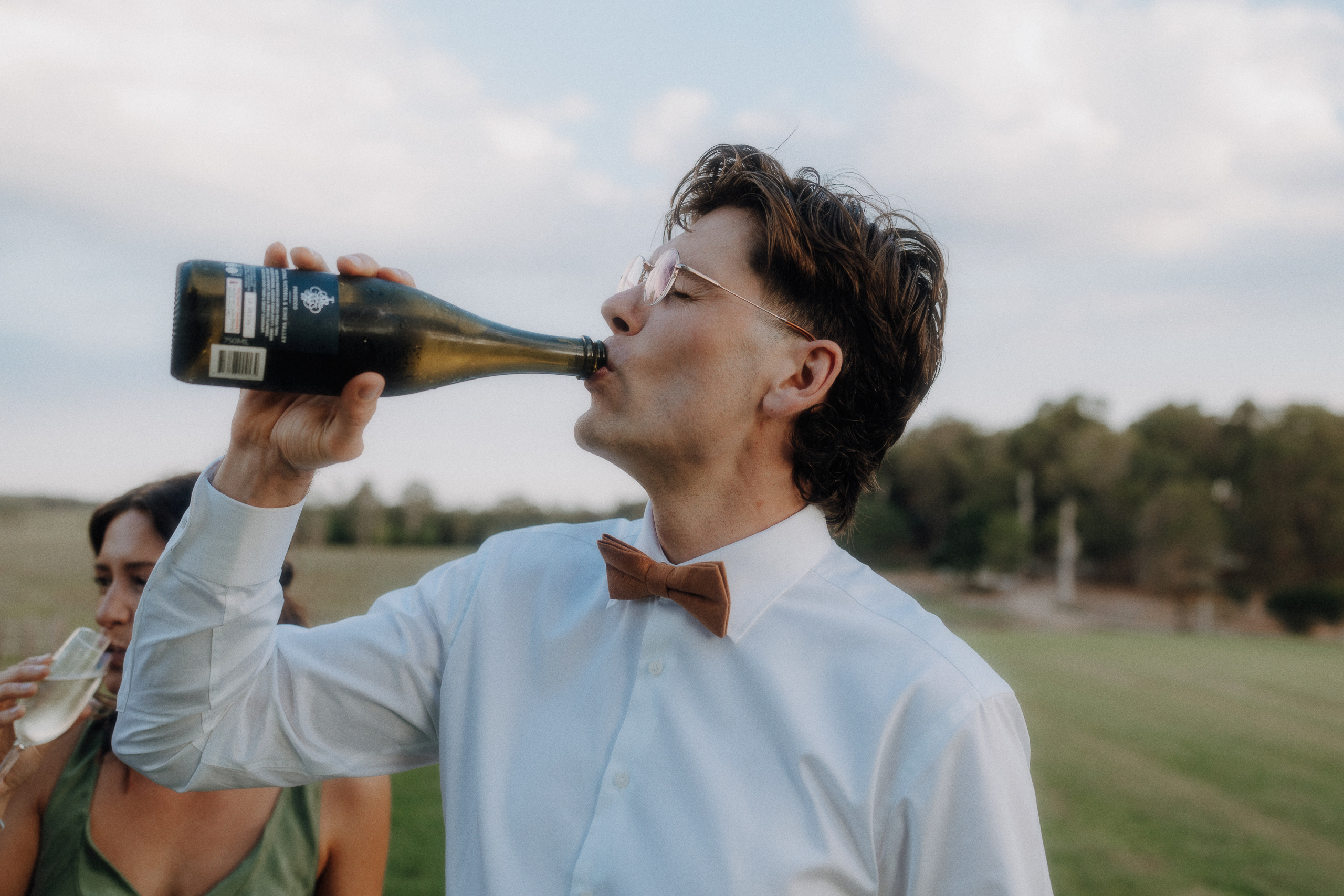 A man in a white shirt and bow tie drinks from a bottle outdoors, with greenery and another person visible in the background.