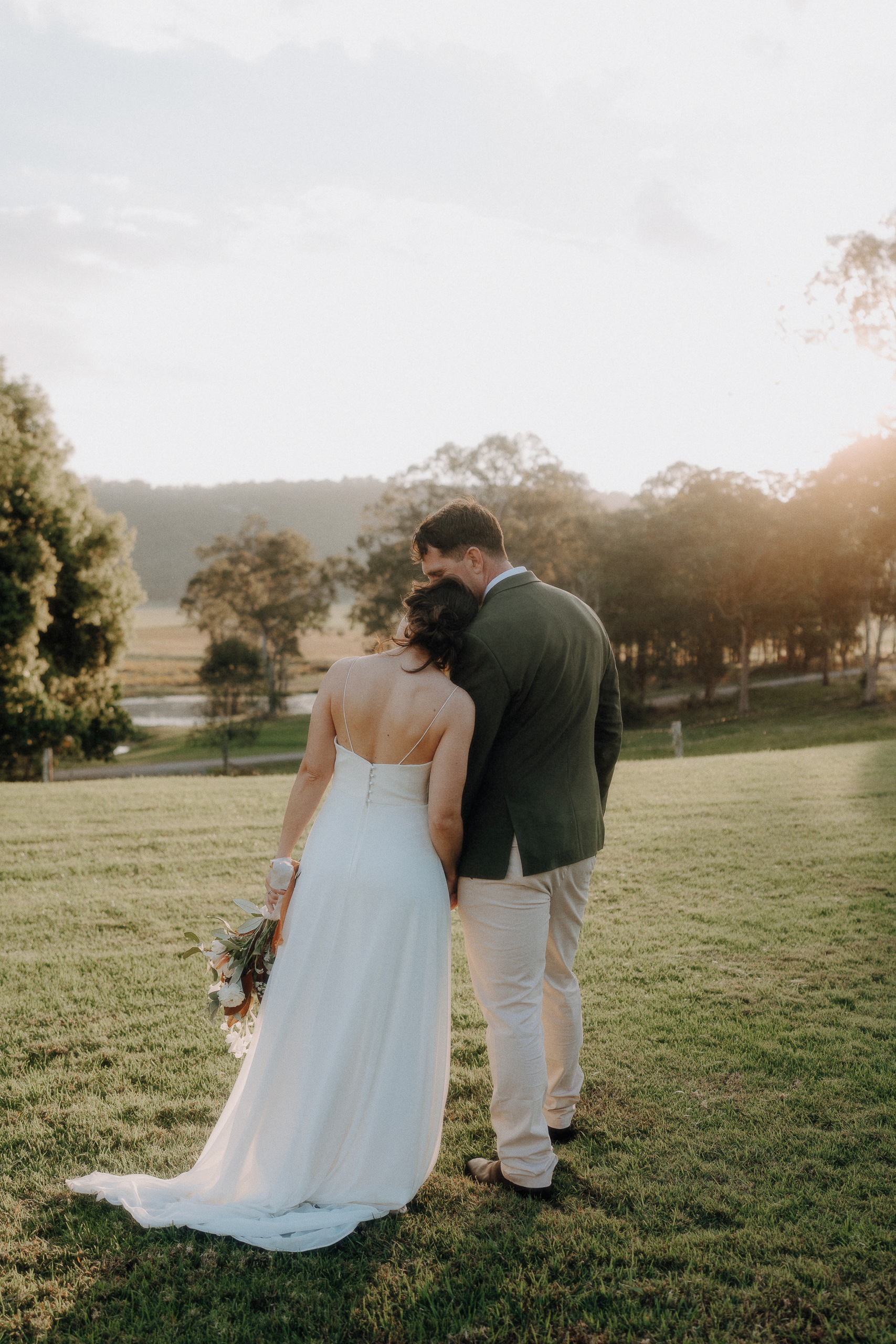 A bride in a white dress leans her head on a groom’s shoulder as they stand together on a grassy field at sunset, holding a bouquet.