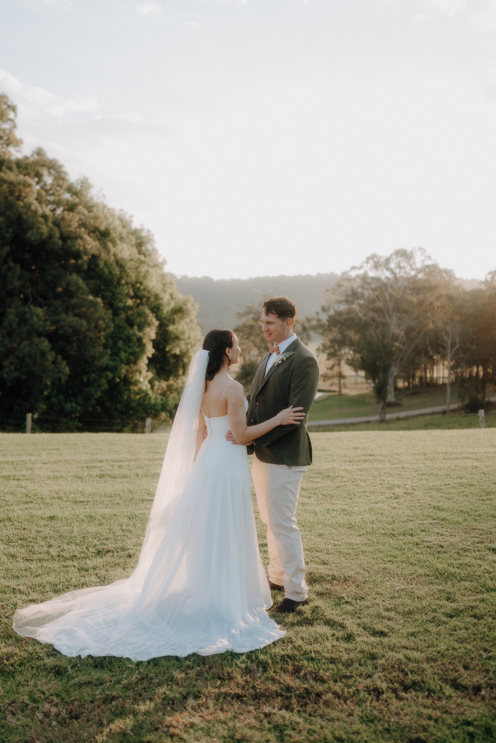 A bride and groom stand facing each other on a grassy field at sunset, surrounded by trees and distant hills.